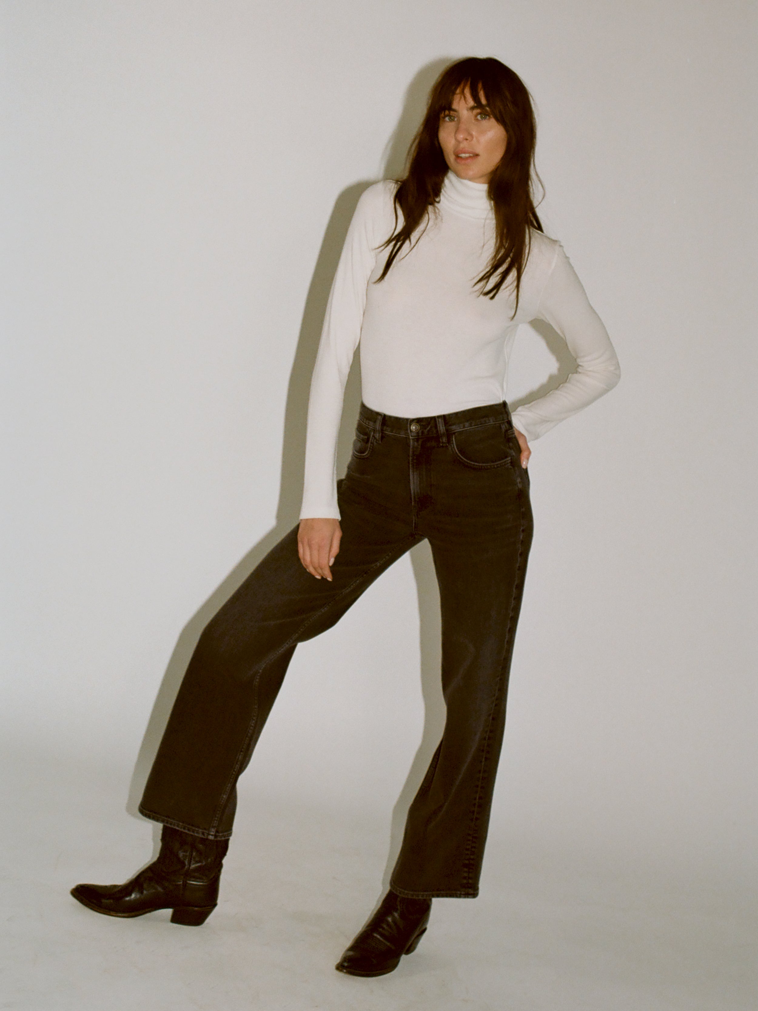 A model wearing a white turtleneck knit with black denim jeans and black boots standing against a plain light gray studio background.