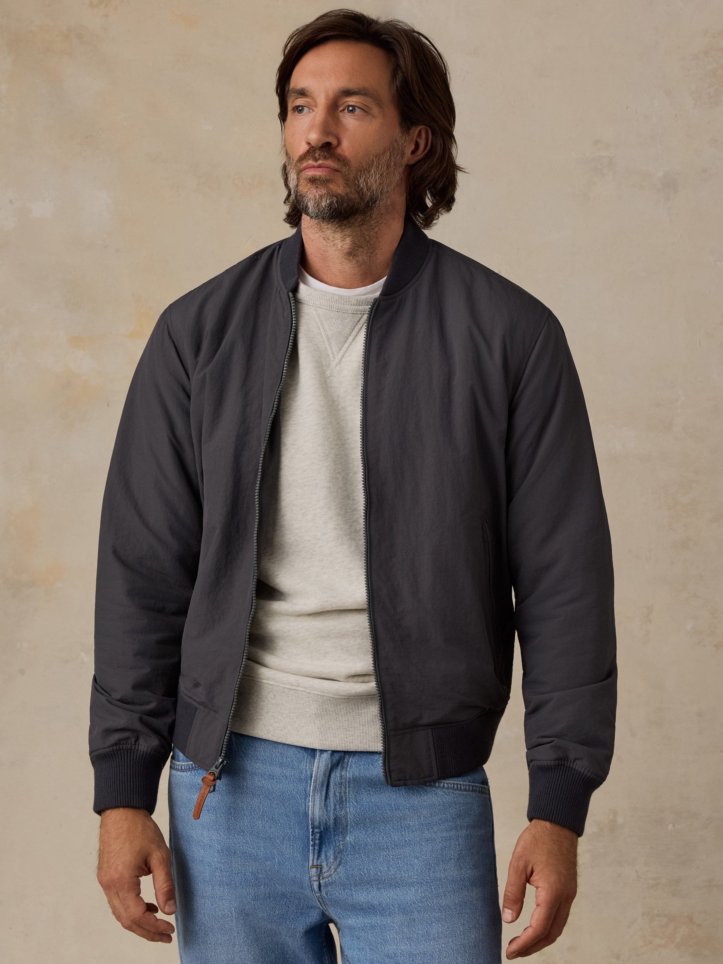 A man wearing a dark charcoal gray bomber jacket over a light gray crewneck sweater and blue jeans, standing against a textured beige wall.