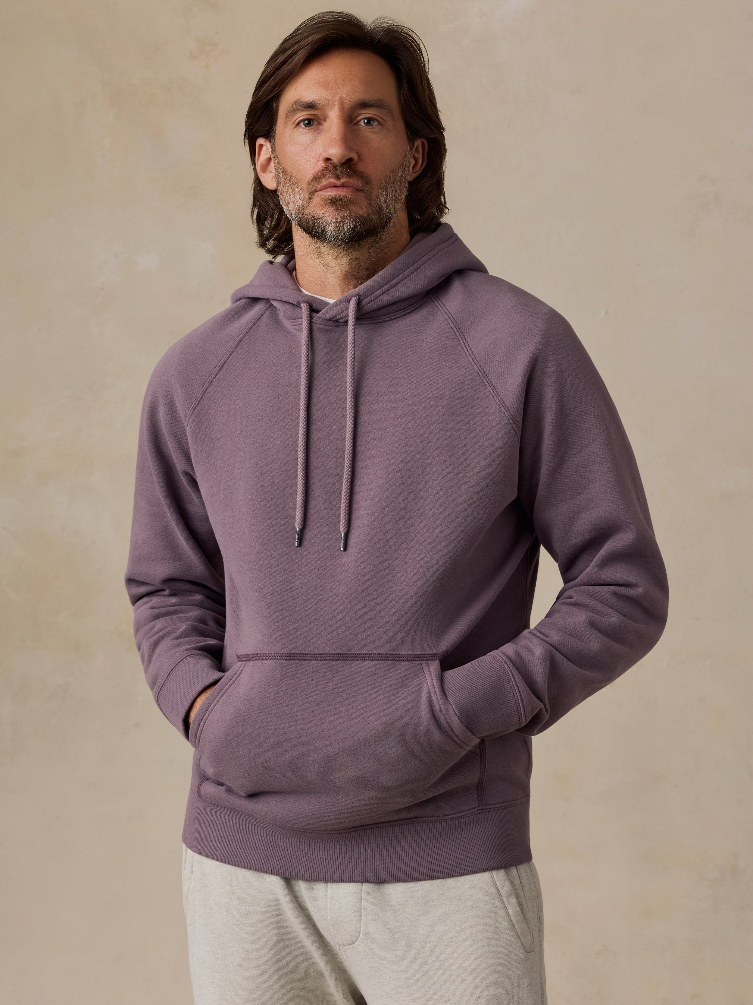 A man wearing a dusty purple pullover hoodie with drawstrings and a front kangaroo pocket, paired with light gray sweatpants, standing against a textured beige wall.