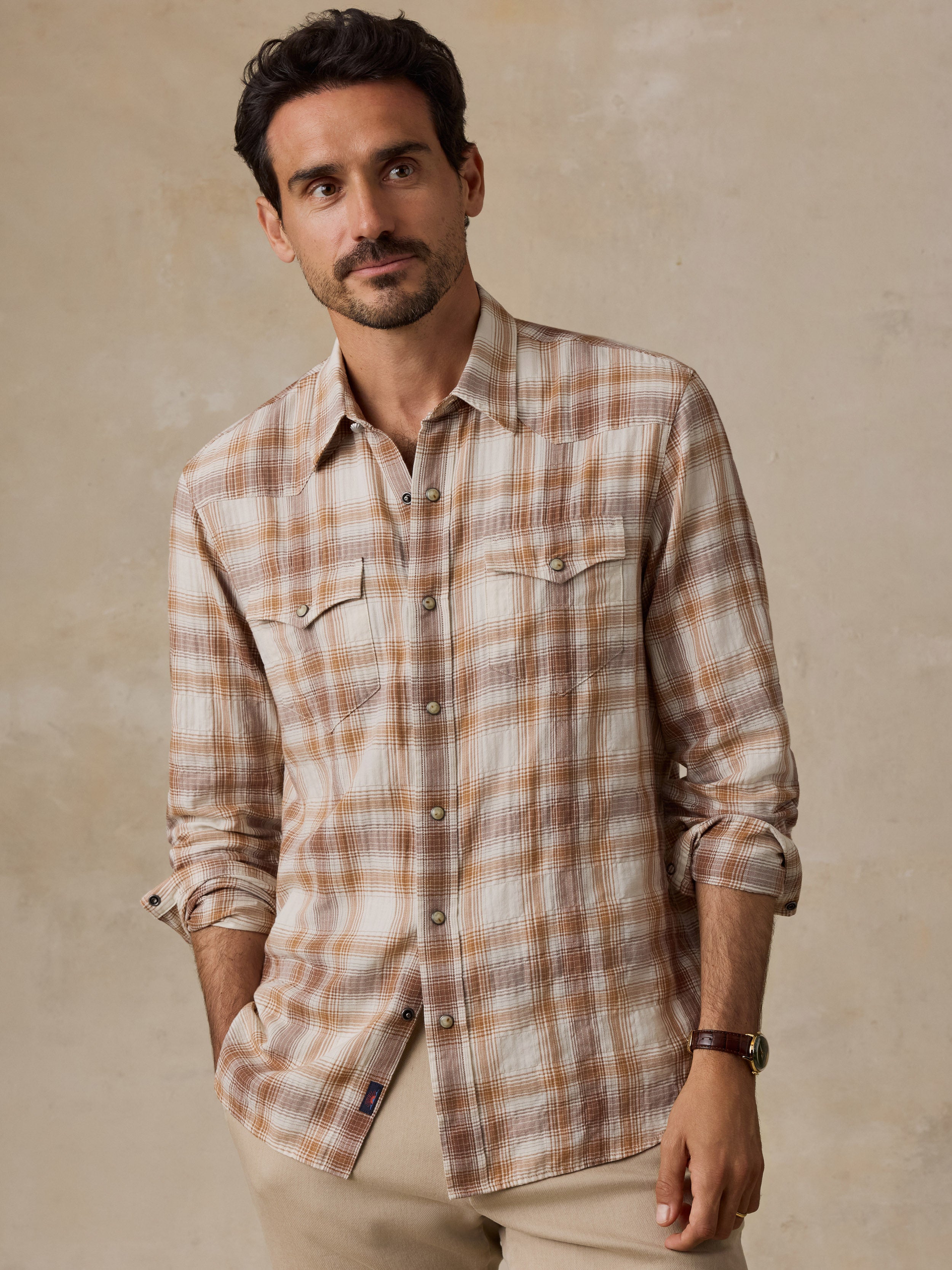 A man wearing a long-sleeve plaid button-up shirt in beige and brown tones, paired with khaki pants, standing against a textured beige background.