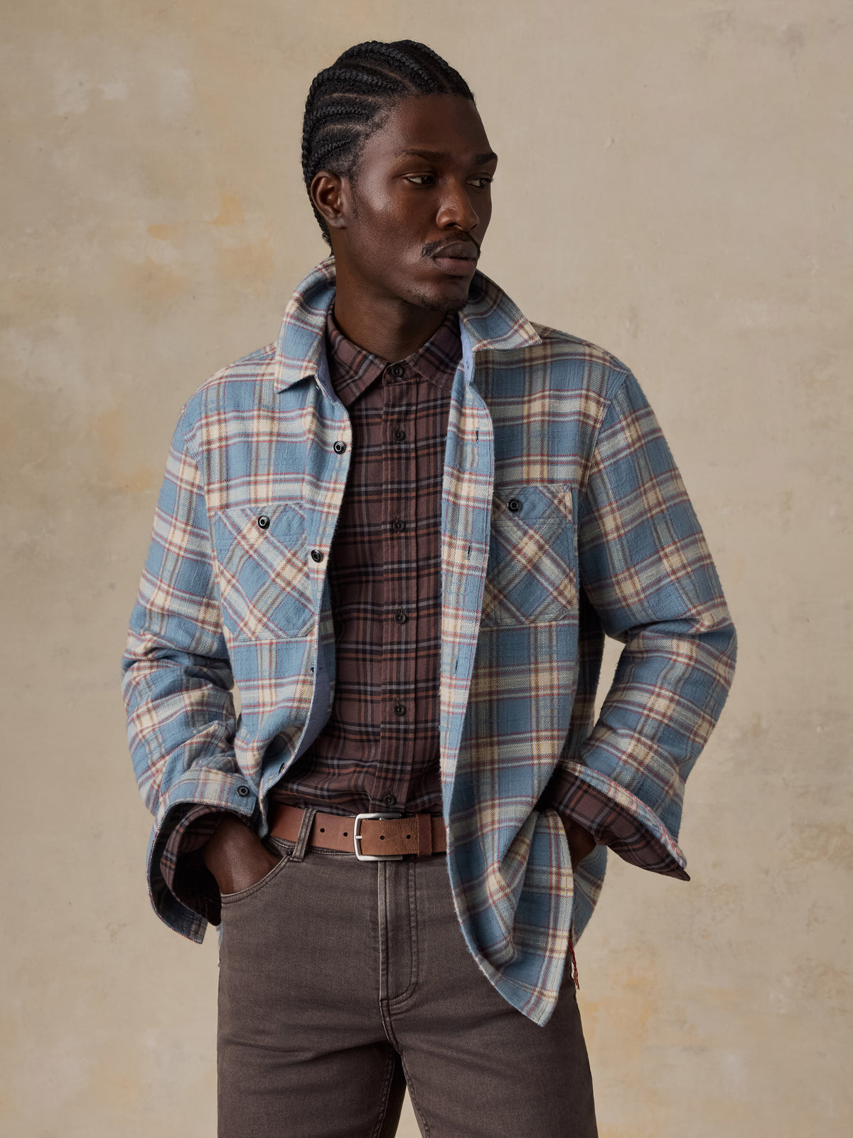 A man wearing a light blue plaid shirt over a plaid button up with brown pants, image 2 of 7