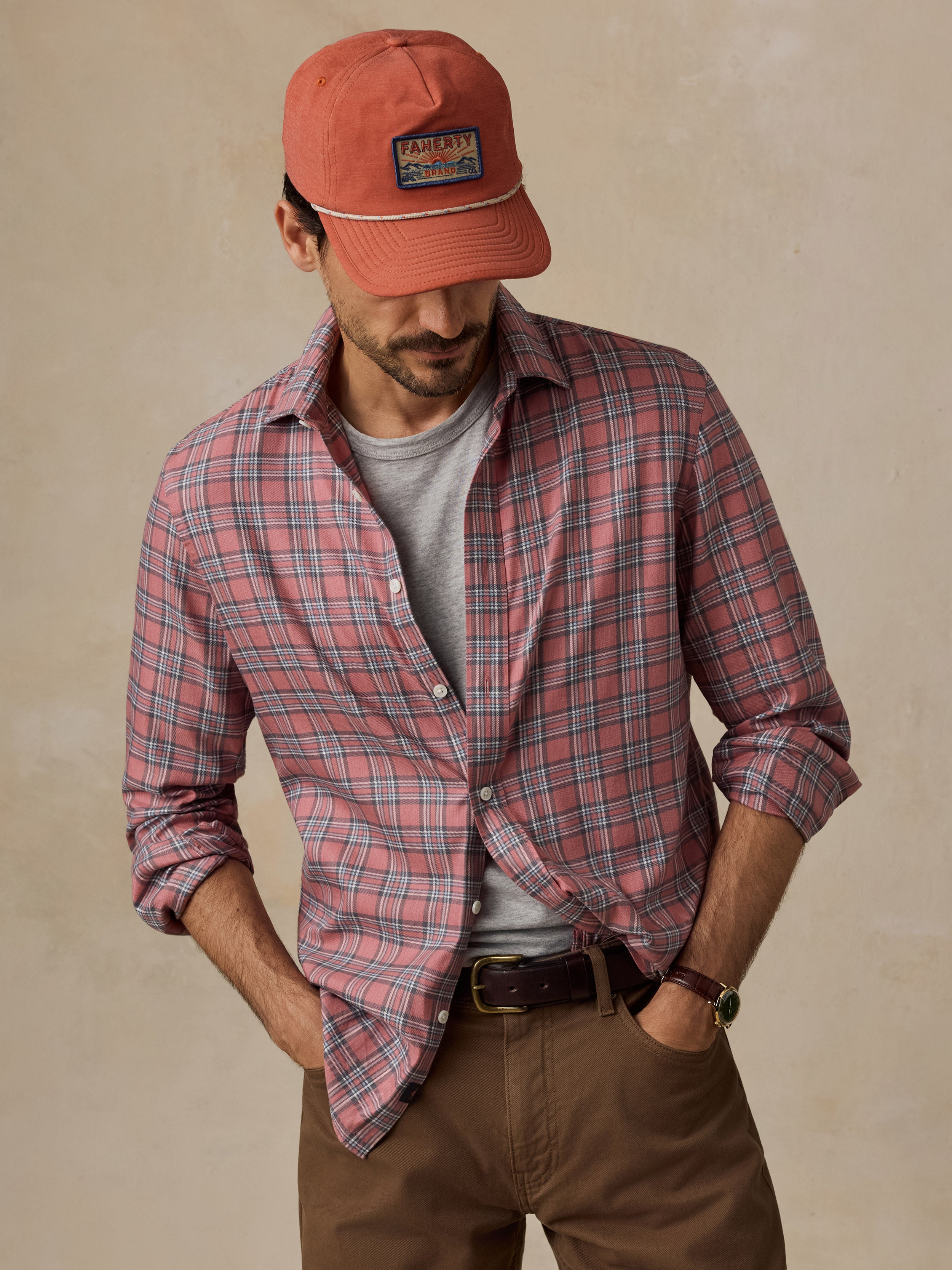 A man wearing an orange baseball cap, a red and blue plaid button-down shirt over a gray t-shirt, brown pants, and a brown belt.