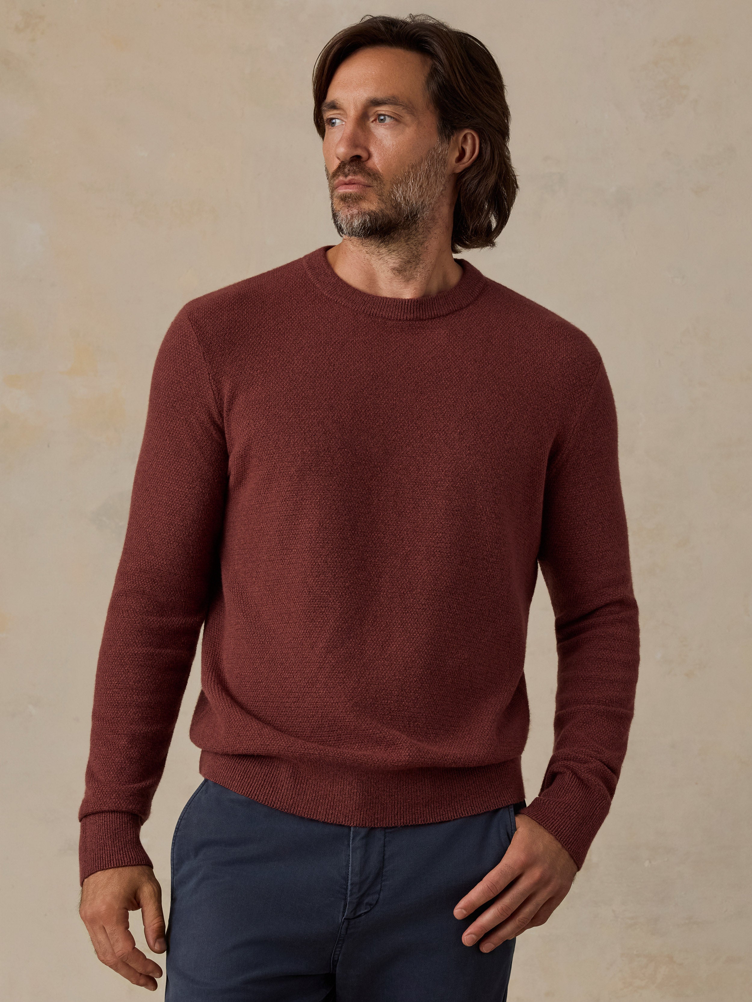 A man wearing a long-sleeve crewneck sweater in a heathered burgundy red, paired with navy blue pants, standing against a textured beige background.