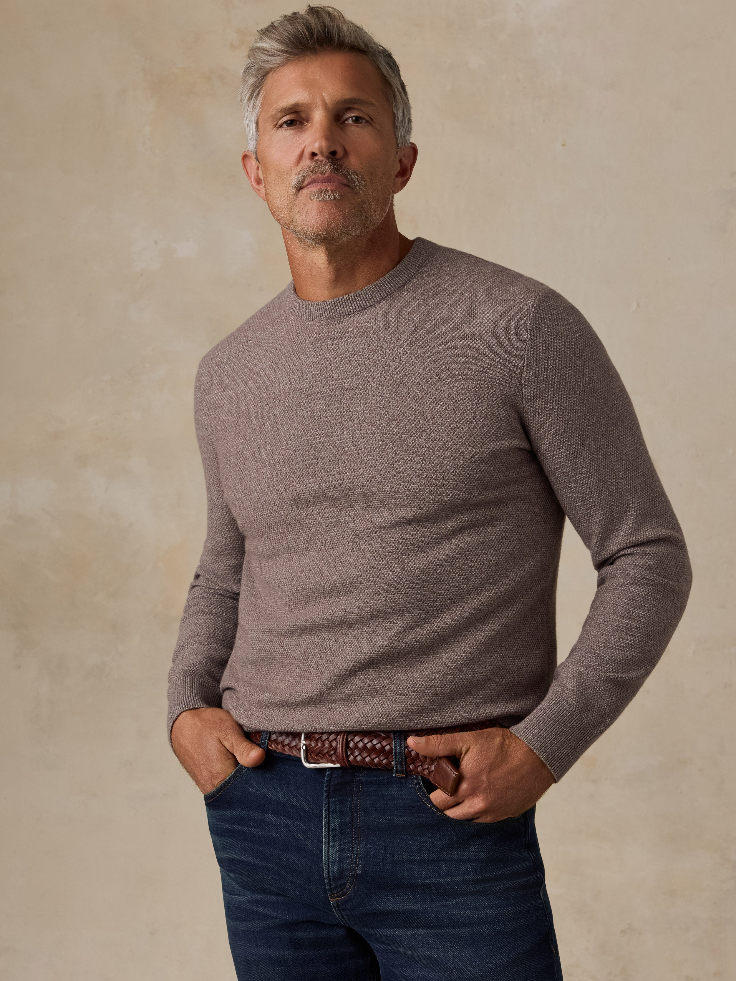 A man wearing a taupe long-sleeve crewneck sweater and dark blue jeans, standing against a textured beige background.