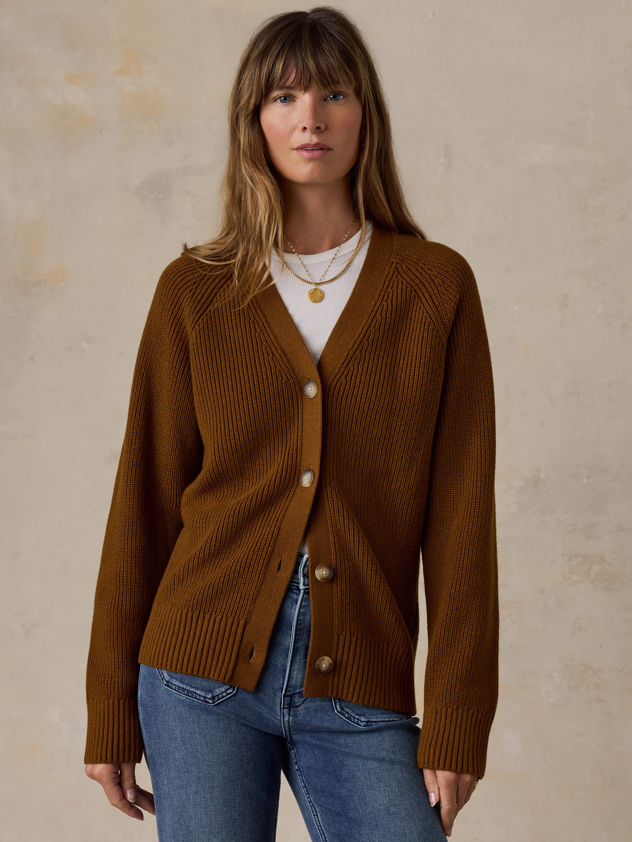 A woman wearing a ribbed brown cardigan over a white top and blue jeans, standing against a textured beige wall.