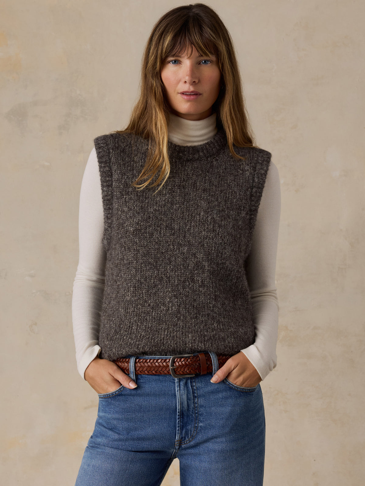 A woman wearing a grey sweater vest with a white turtleneck and blue jeans, image 1 of 5