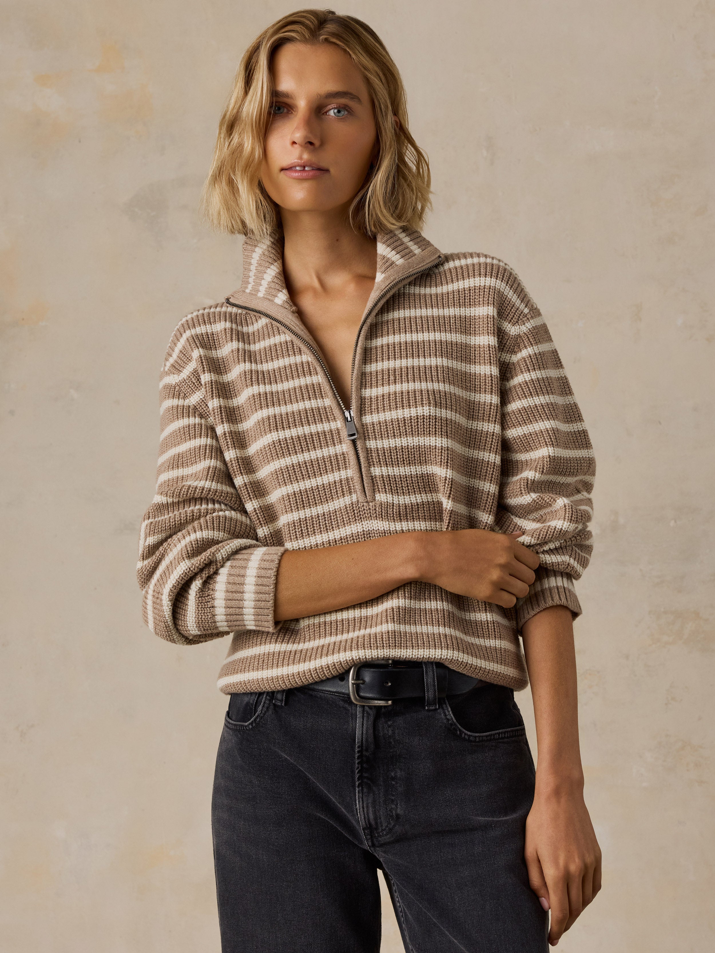 A woman wearing a beige and brown striped quarter-zip pullover sweater with dark wash jeans