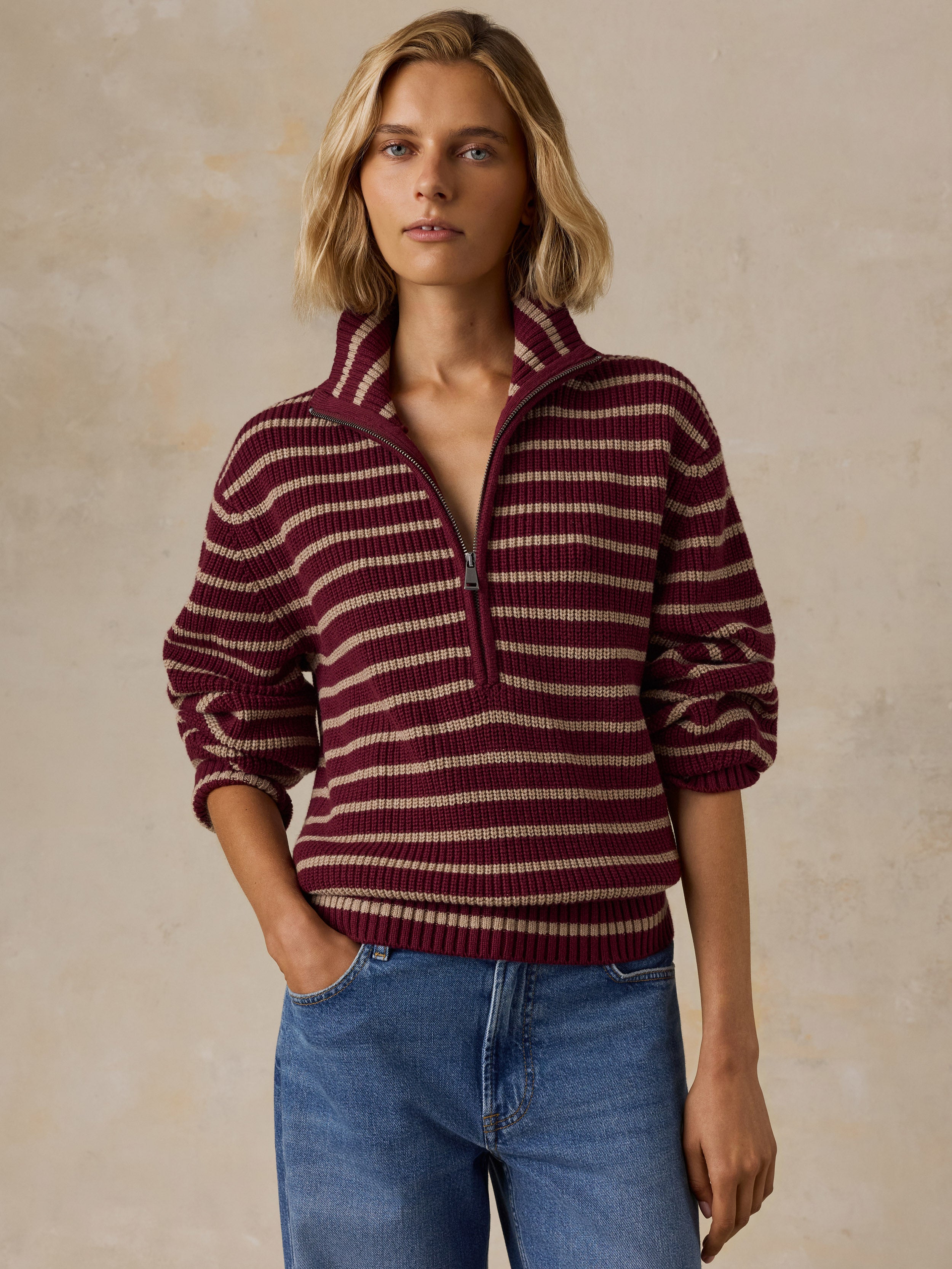 A woman wearing a burgundy and beige striped quarter-zip knit sweater with blue jeans