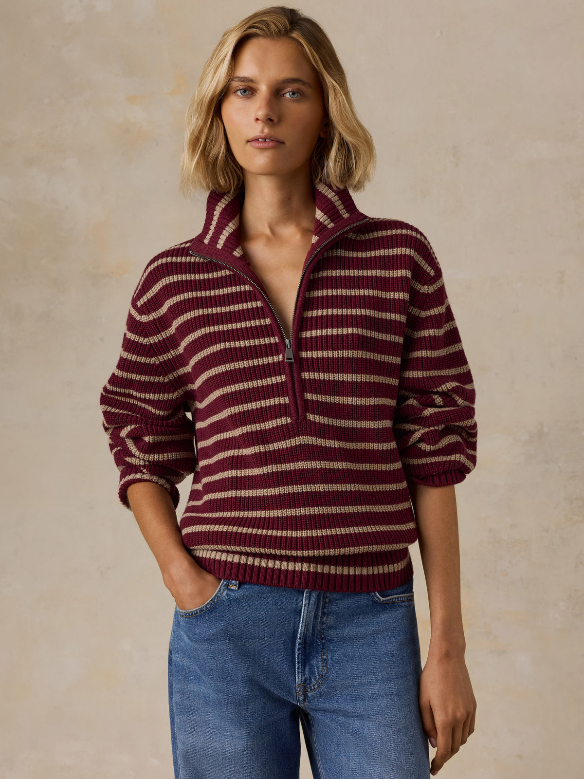 A woman wearing a burgundy and beige striped quarter-zip knit sweater with blue jeans, image 1 of 5
