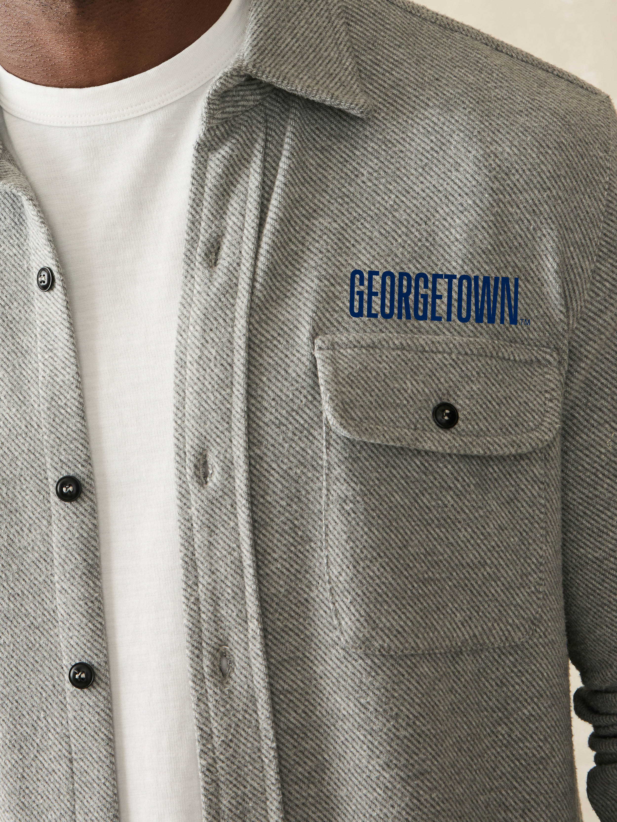 Man wearing a grey long sleeve button up shirt with Georgetown graphic on the left chest and left arm against a beige background