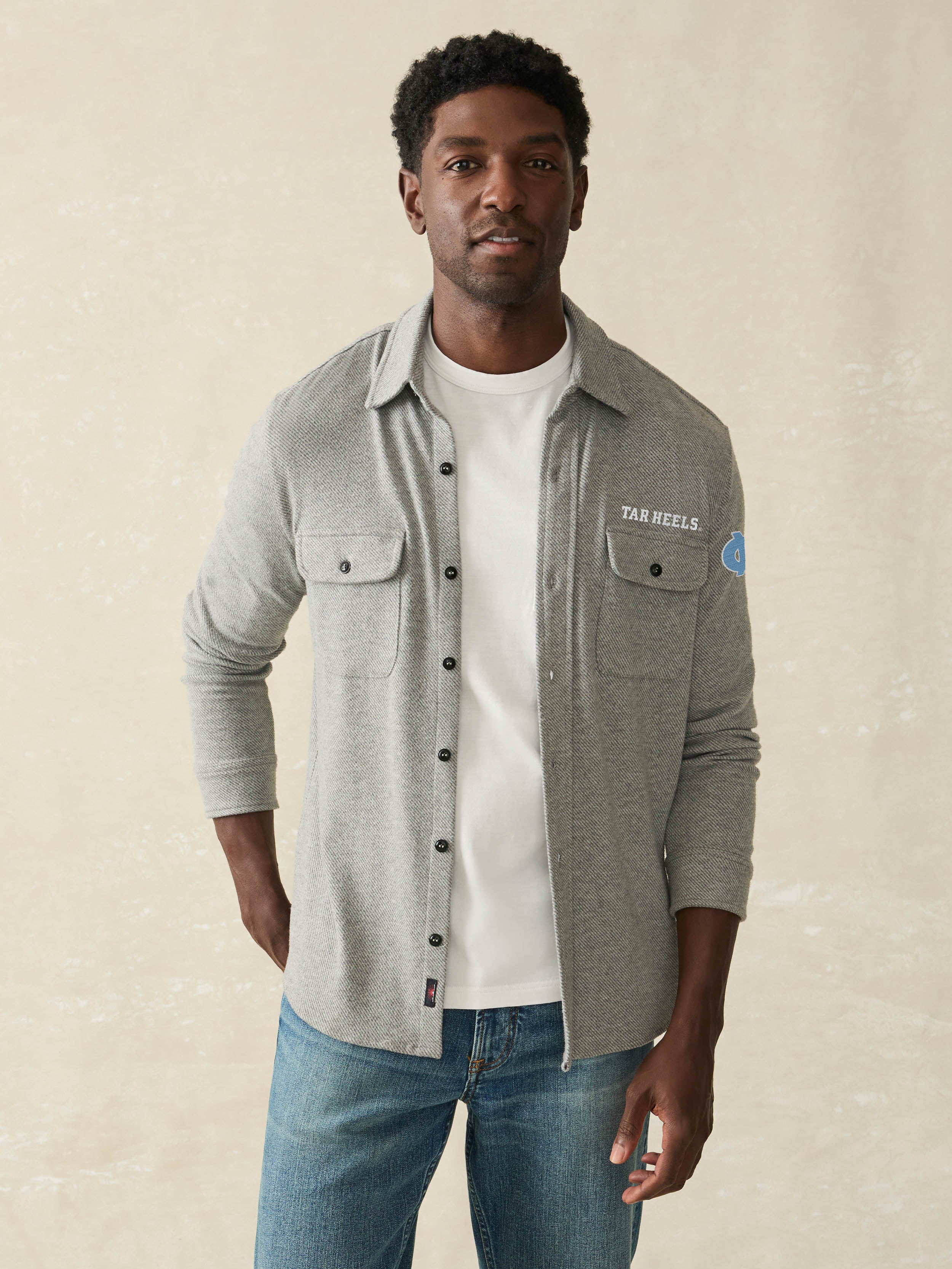 Man wearing a grey long sleeve button up shirt with UNC graphic at the left chest and left arm against a beige background