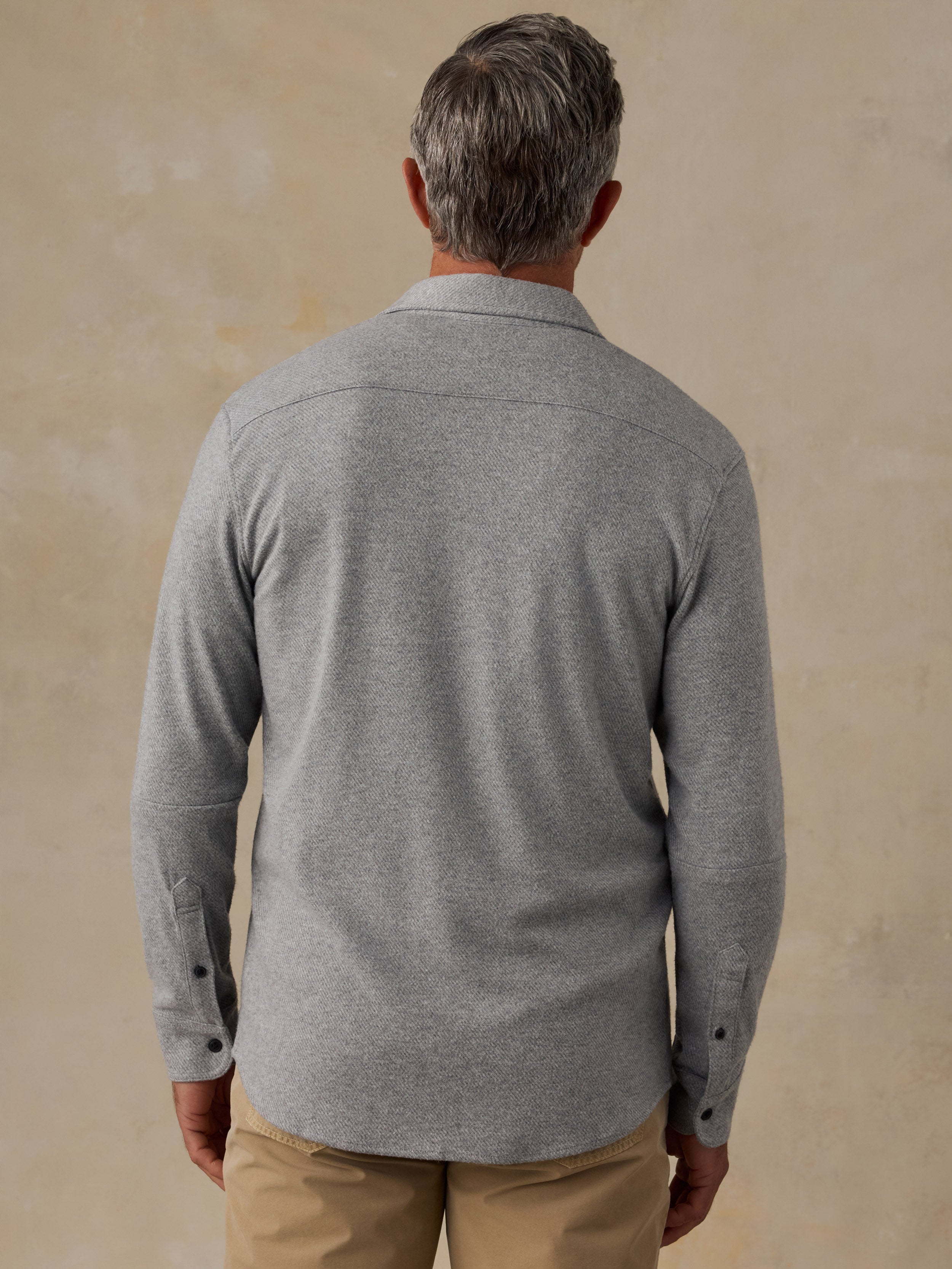 A man viewed from behind wearing a light gray long-sleeve knit shirt and khaki pants, standing against a textured beige wall.