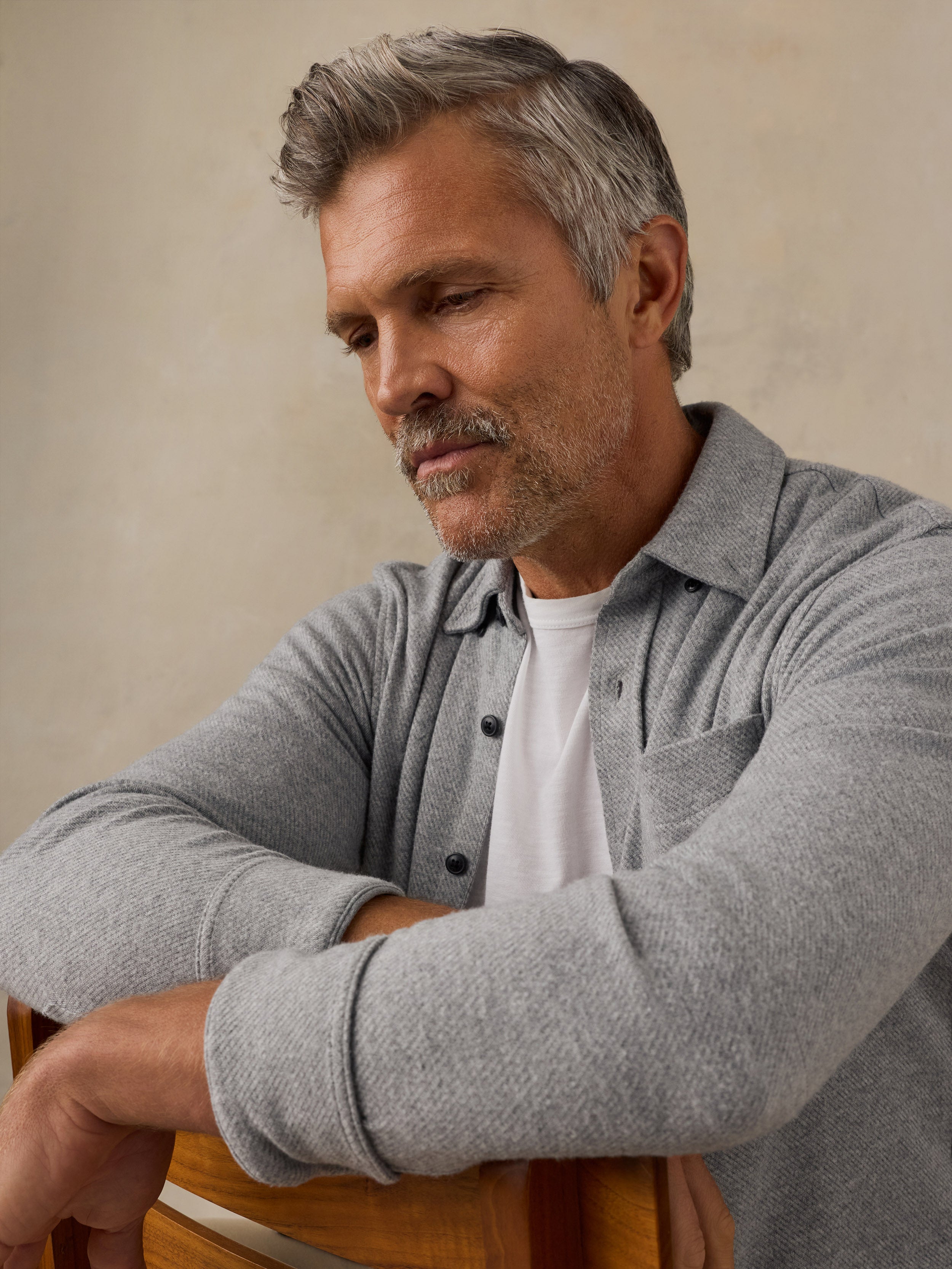 A man with gray hair and a mustache wearing a light gray long-sleeve button-up shirt over a white t-shirt, leaning on a wooden chair against a textured beige wall.
