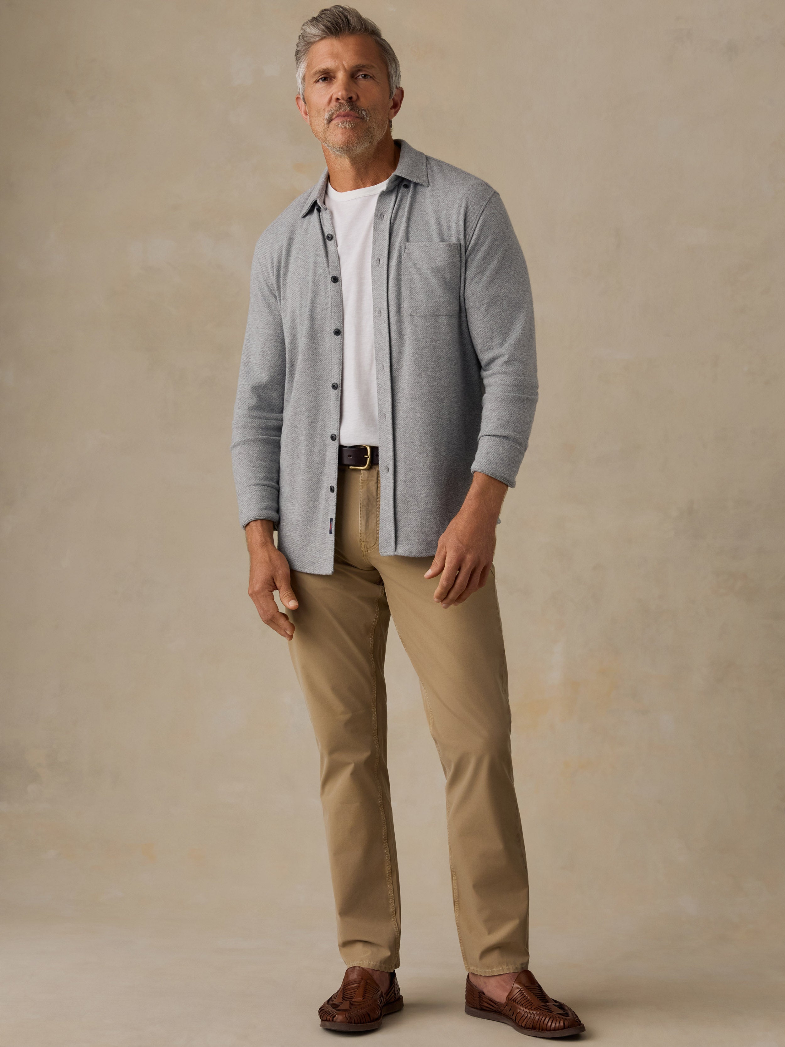 A man wearing a light gray button-down shirt over a white t-shirt, khaki pants, and brown leather loafers, standing against a textured beige studio backdrop.