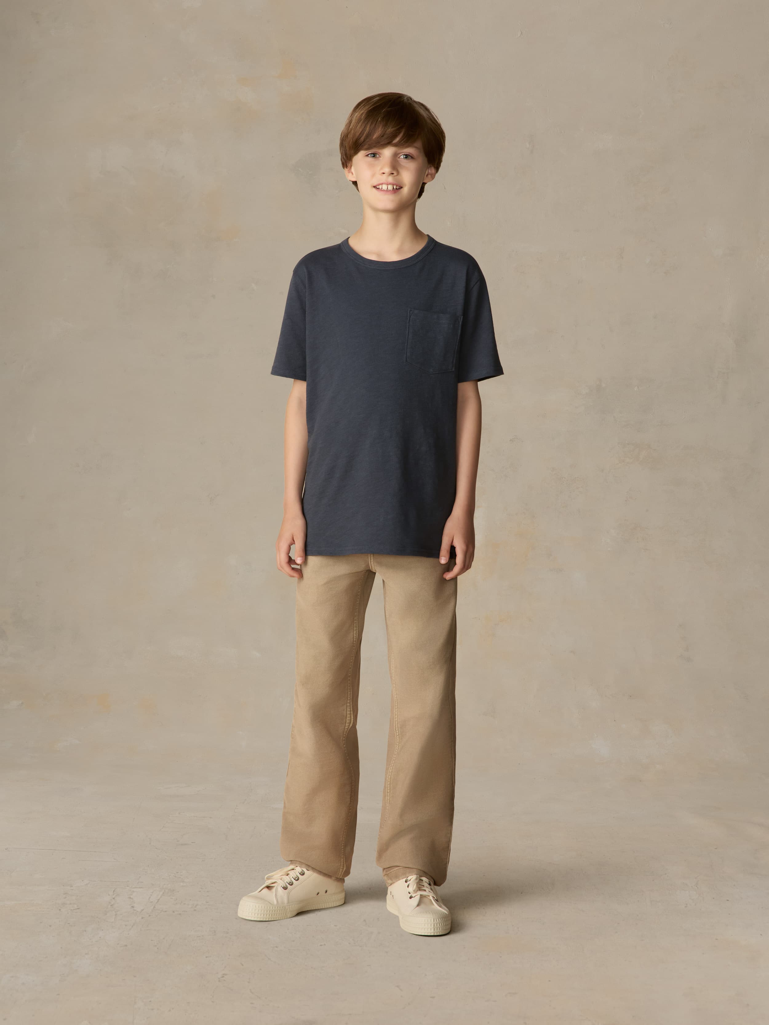 Child wearing a blue pocket tee and beige pants against a beige background