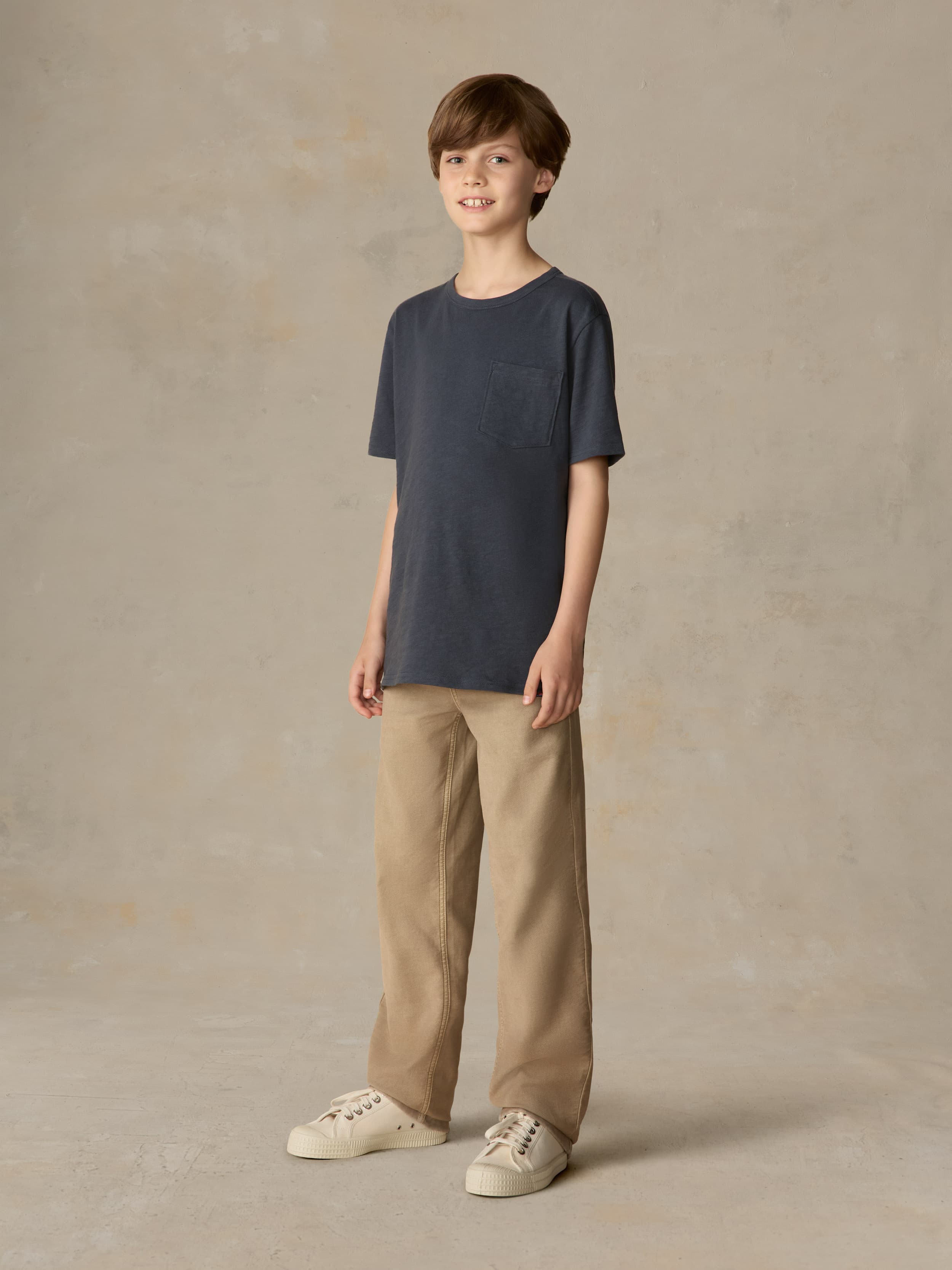 Child wearing a blue pocket tee and beige pants against a beige background