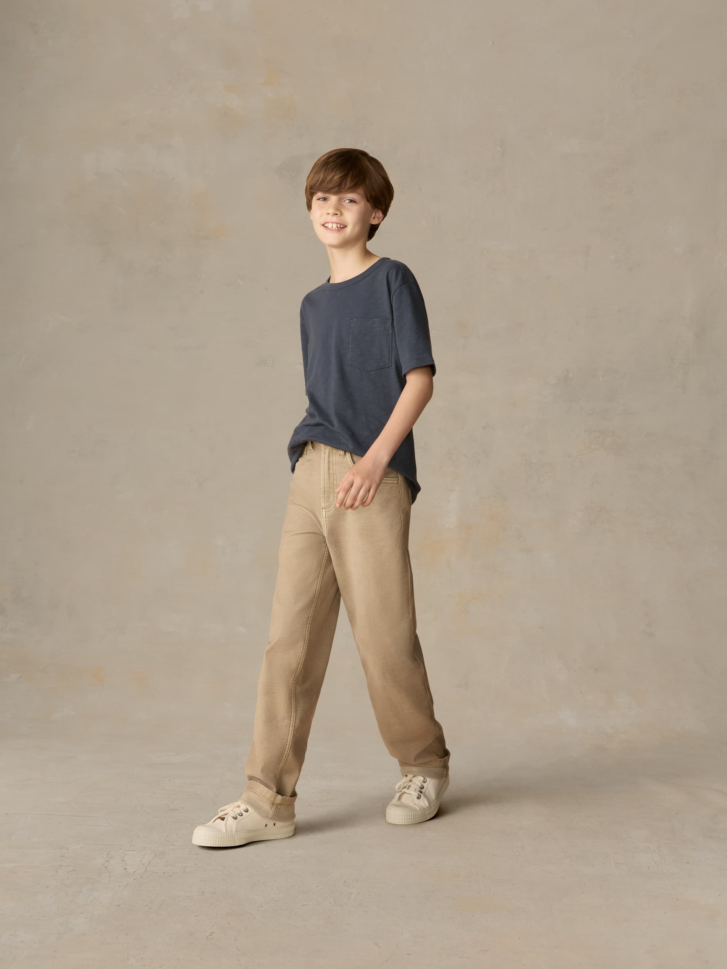 Child wearing a blue pocket tee and beige pants against a beige background