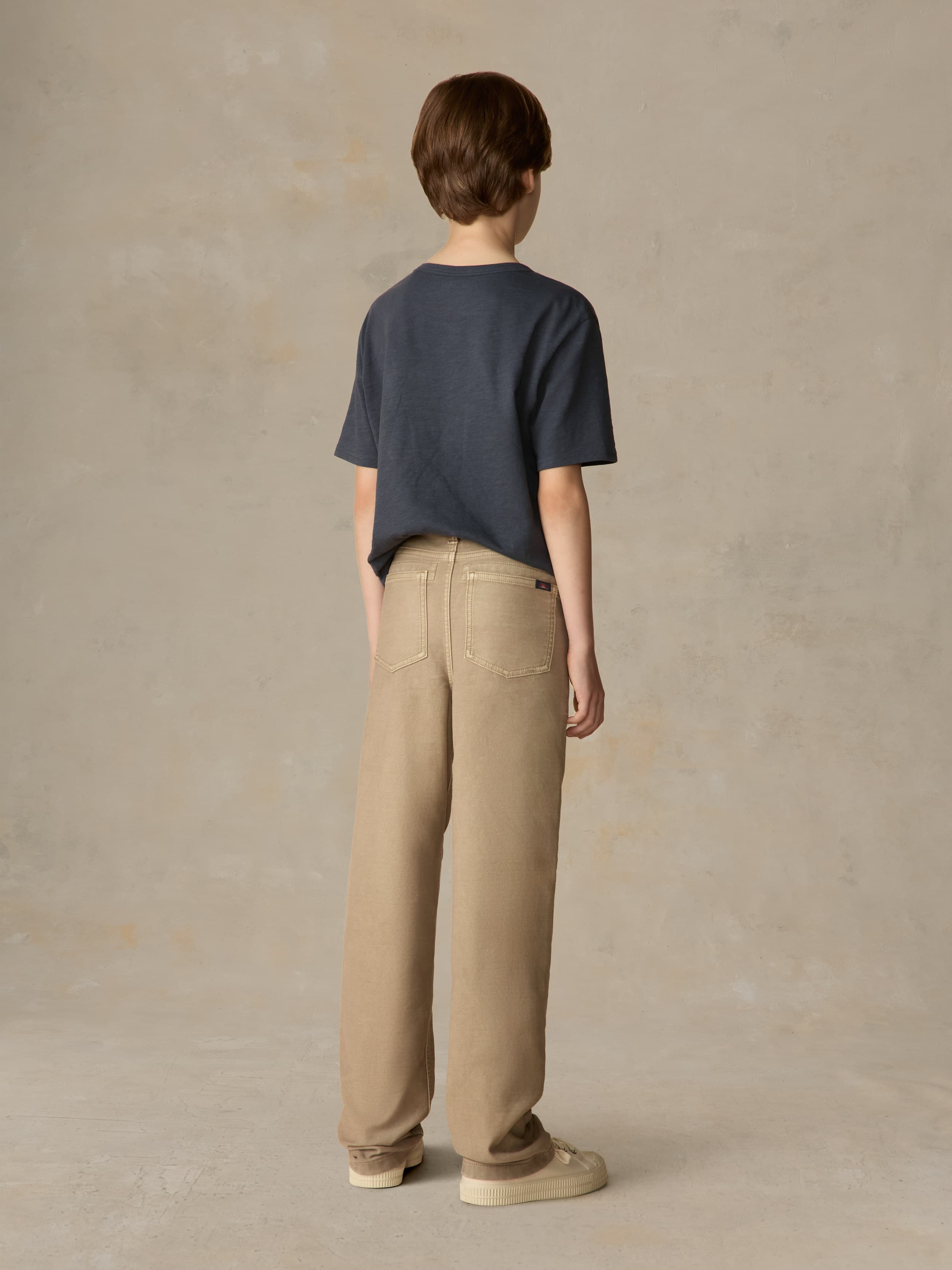 Child wearing a blue pocket tee and beige pants against a beige background