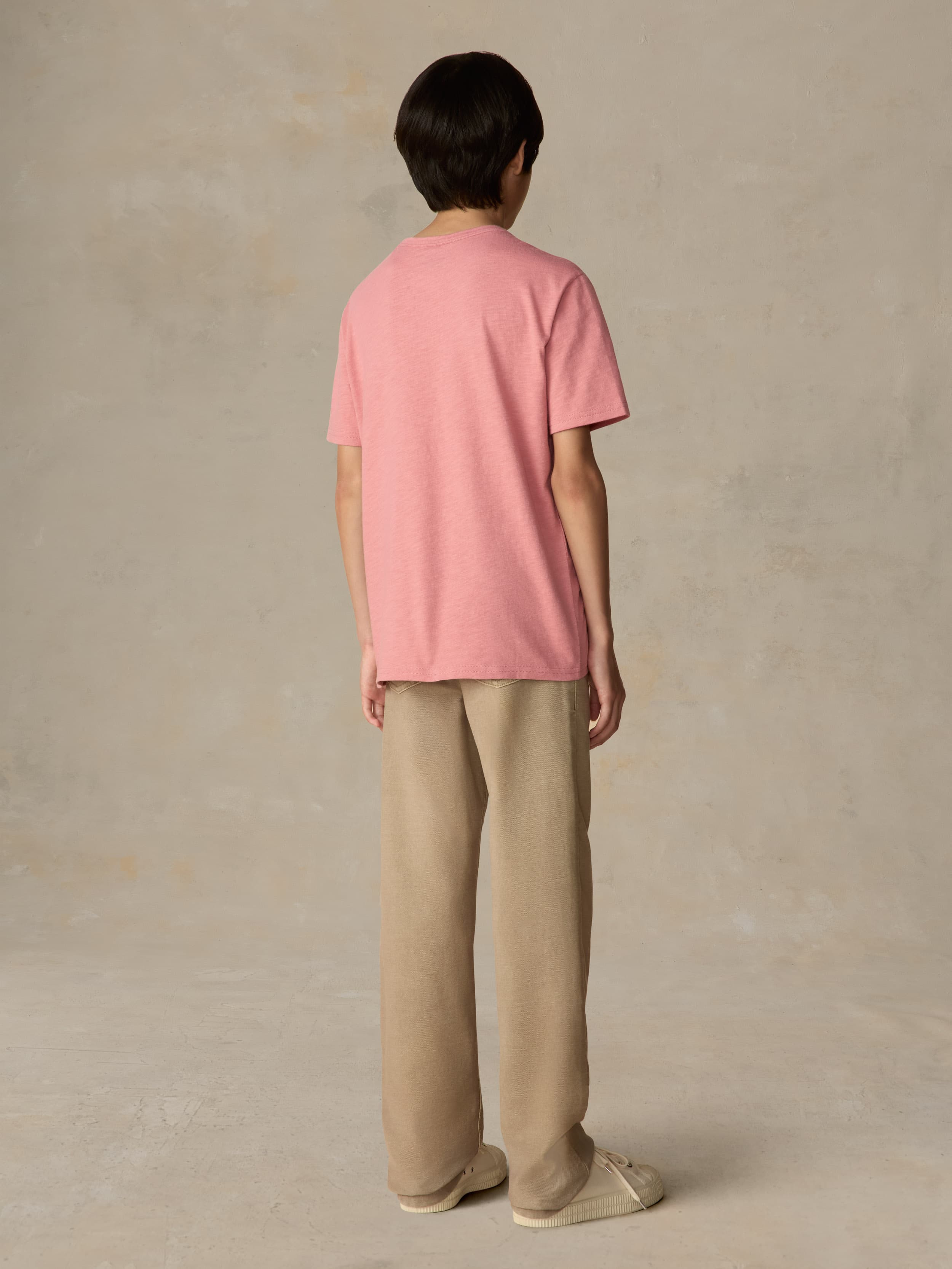 Child wearing a red pocket tee and beige pants against a beige background