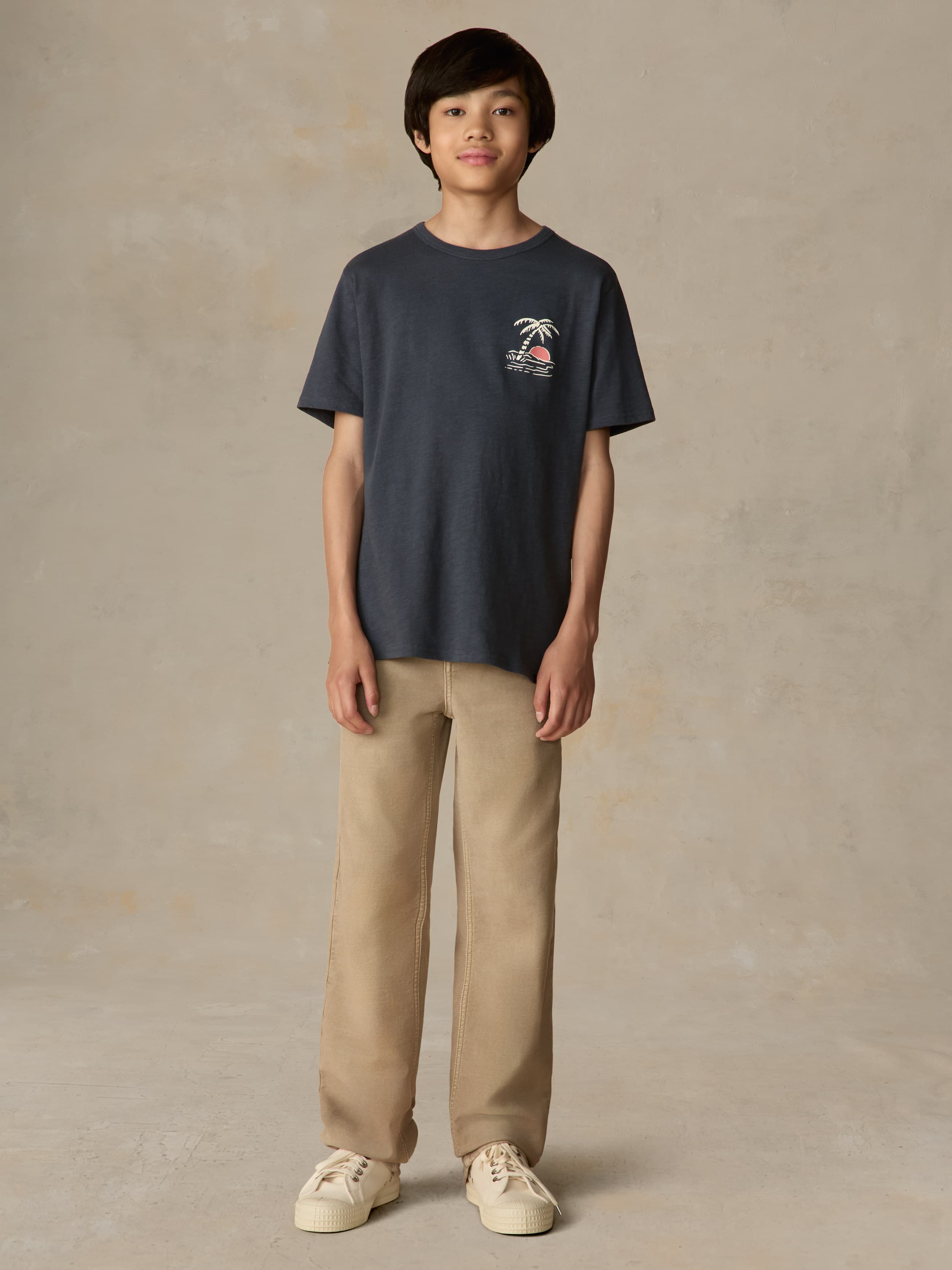 Child wearing a blue graphic shirt and beige pants against a beige background