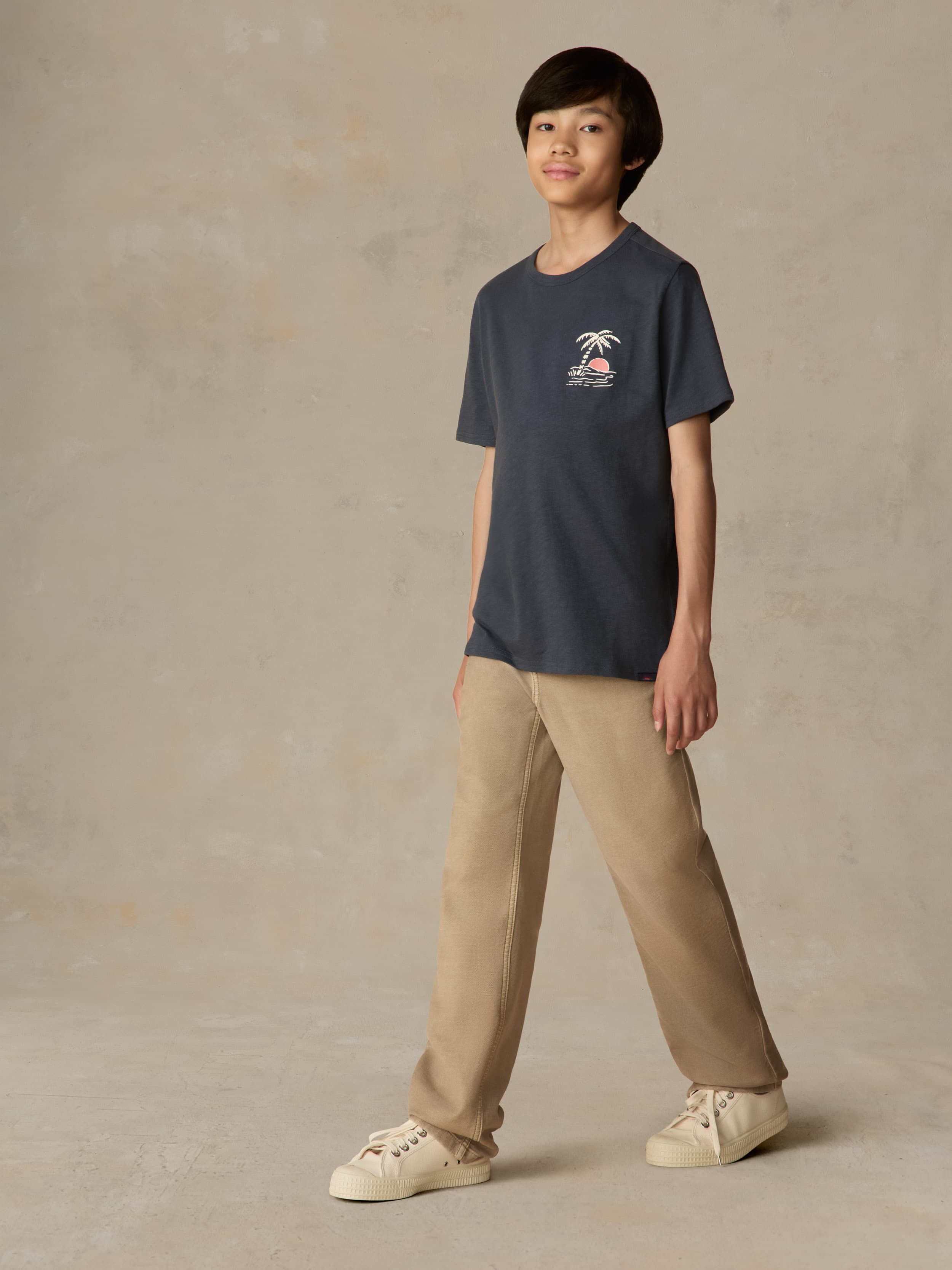 Child wearing a blue graphic shirt and beige pants against a beige background