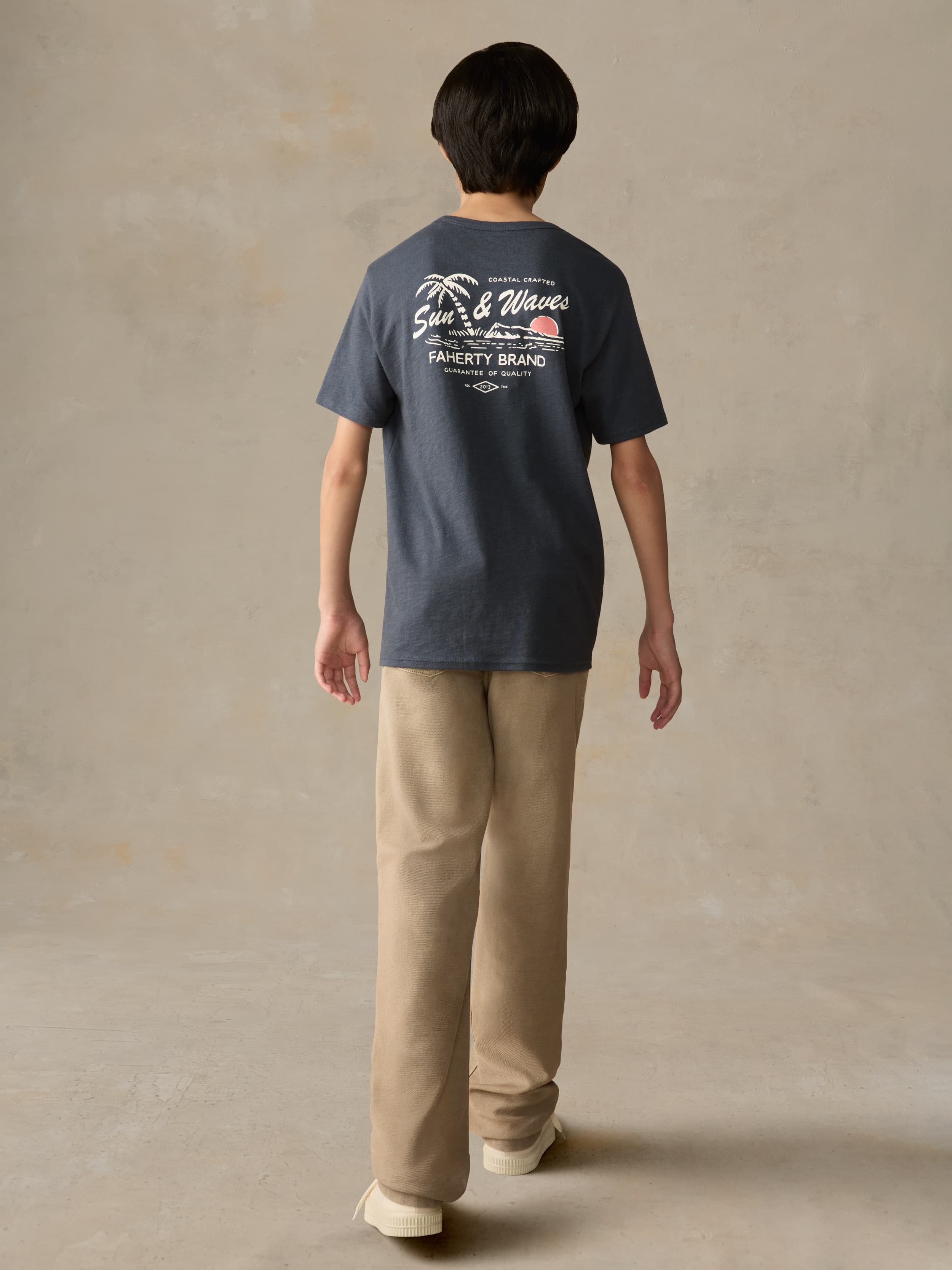 Child wearing a blue graphic shirt and beige pants against a beige background