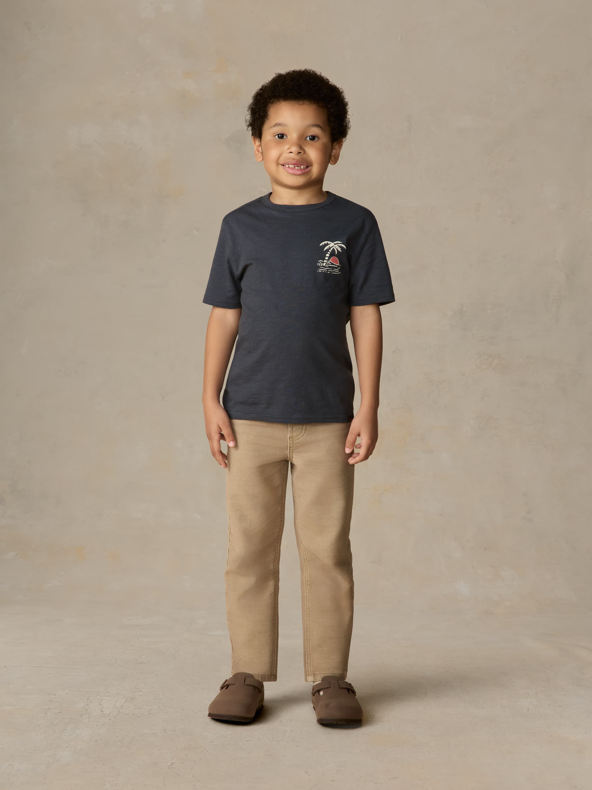 Child wearing a blue graphic shirt and beige pants against a beige background