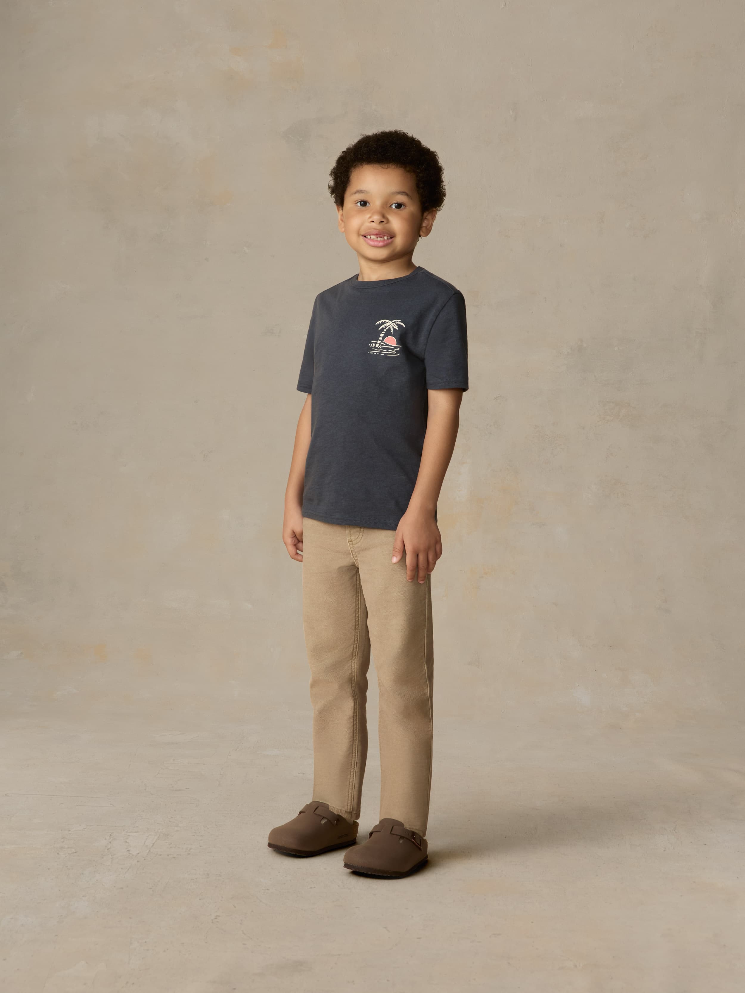 Child wearing a blue graphic shirt and beige pants against a beige background