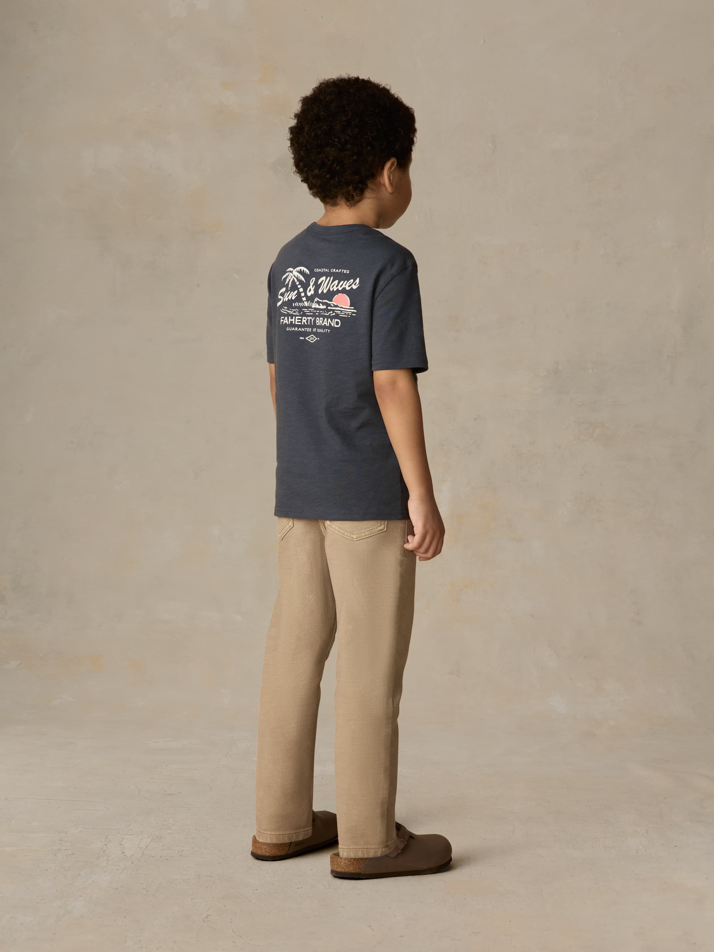 Child wearing a blue graphic shirt and beige pants against a beige background
