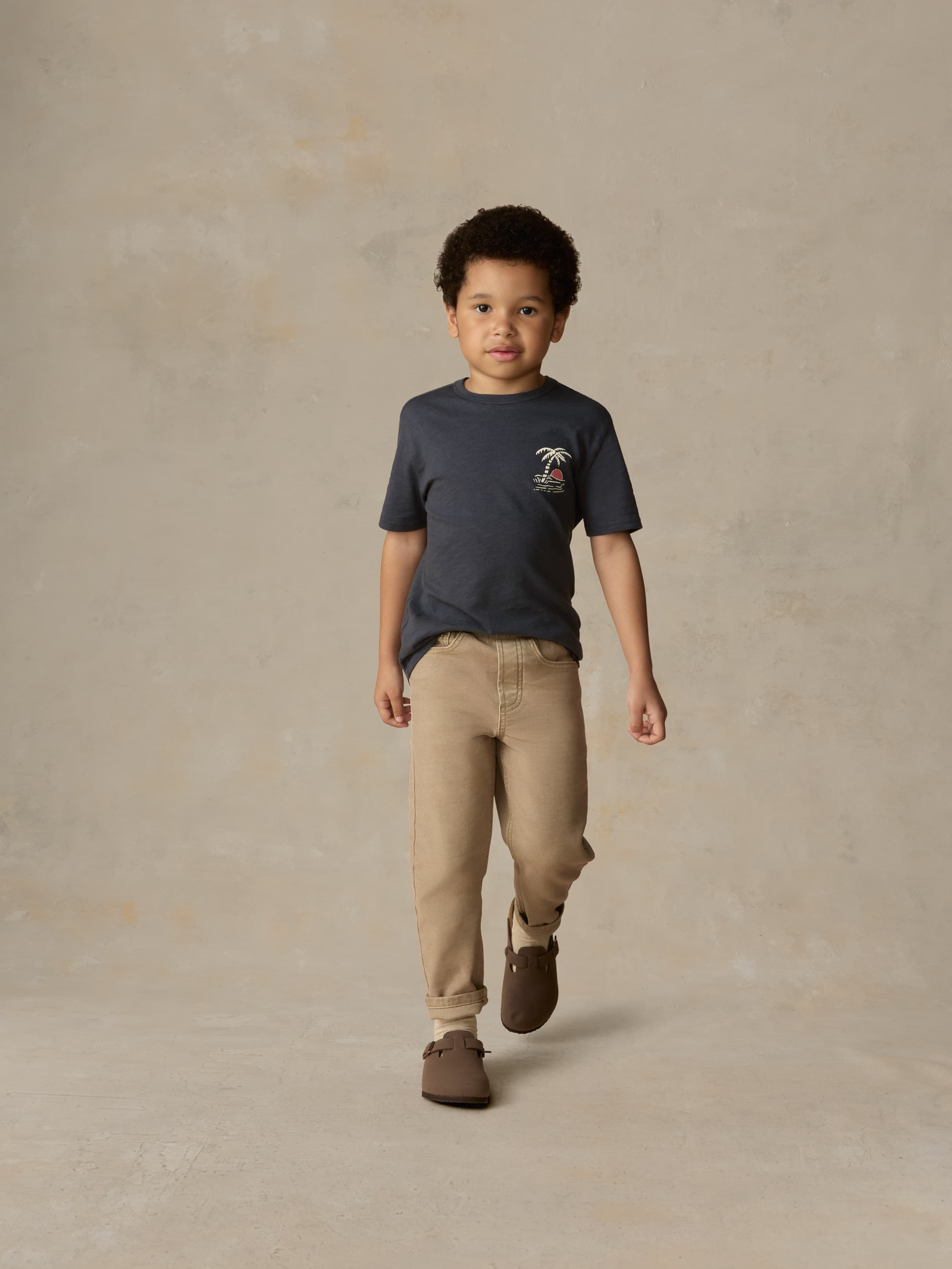 Child wearing a blue graphic shirt and beige pants against a beige background