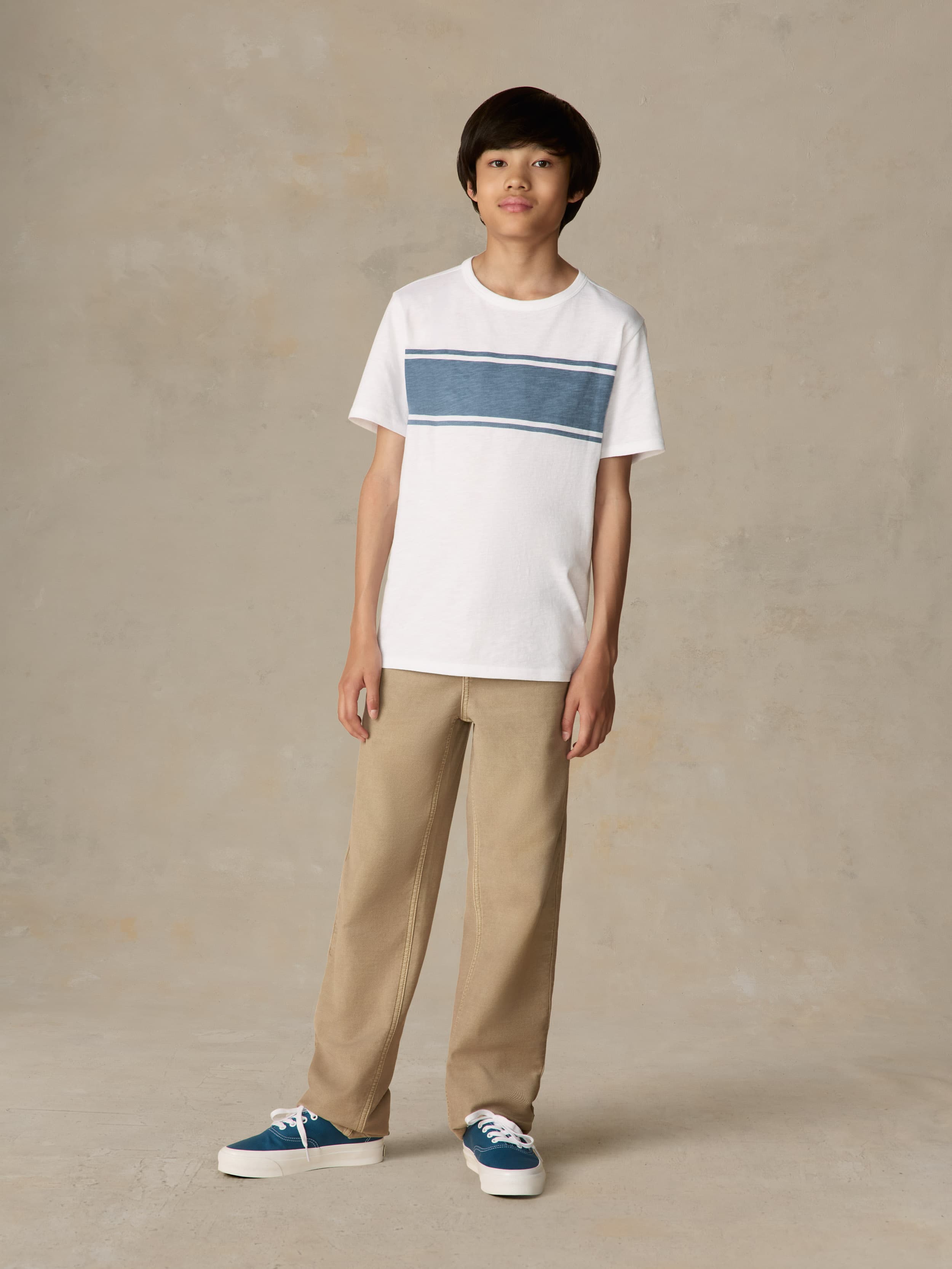 Child wearing a white shirt with blue stripe and beige pants against a beige background