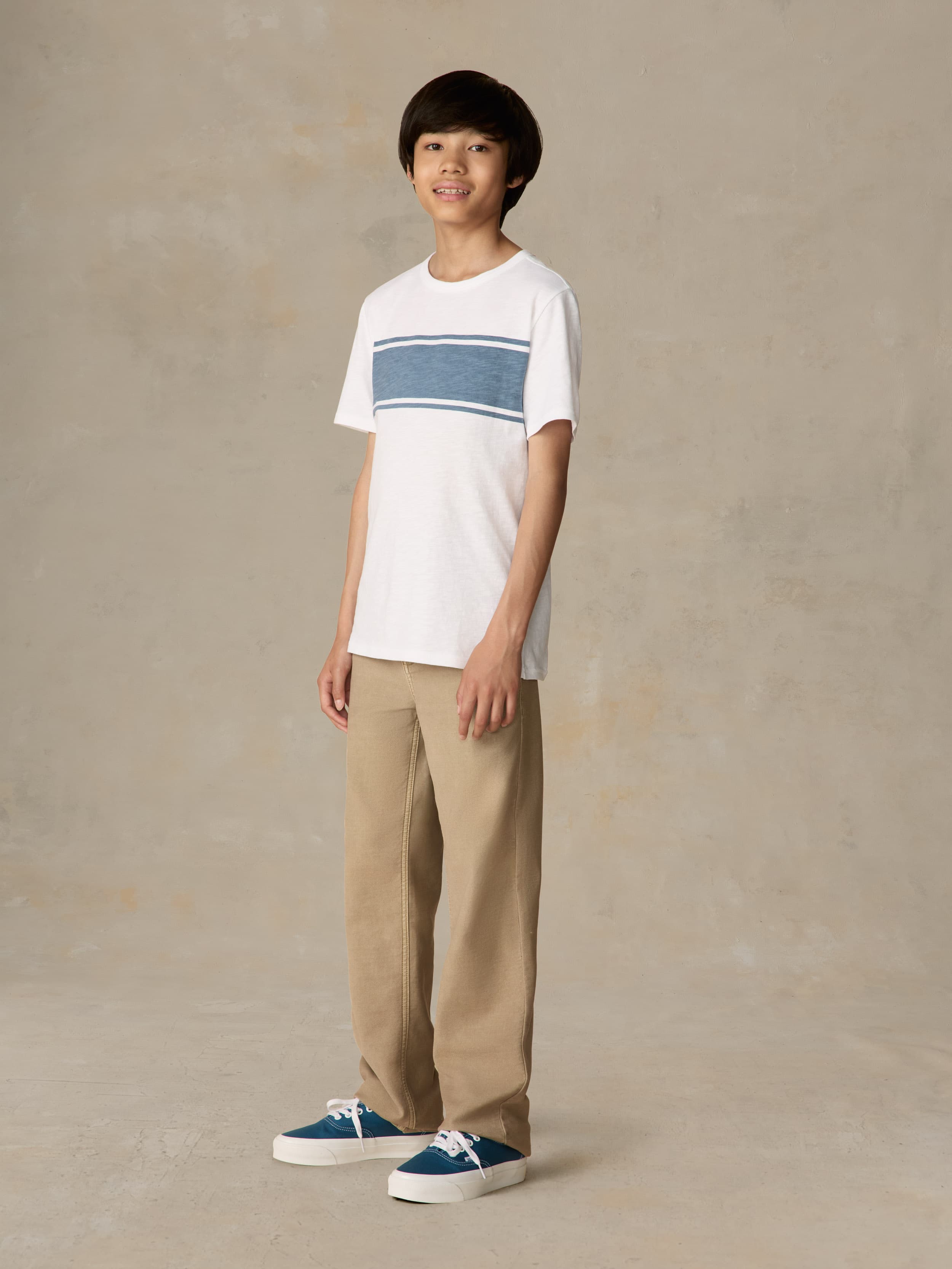 Child wearing a white shirt with blue stripe and beige pants against a beige background
