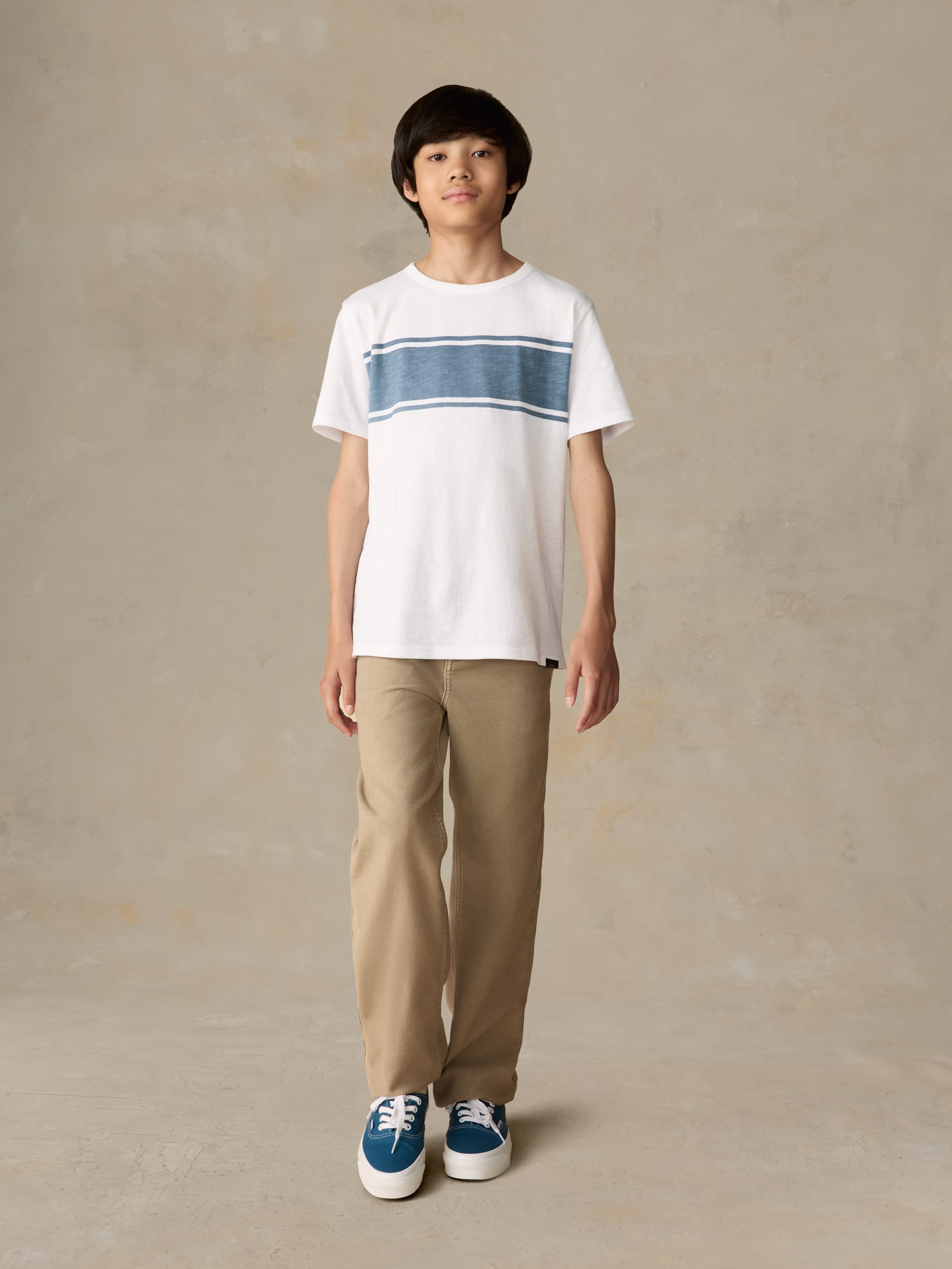 Child wearing a white shirt with blue stripe and beige pants against a beige background