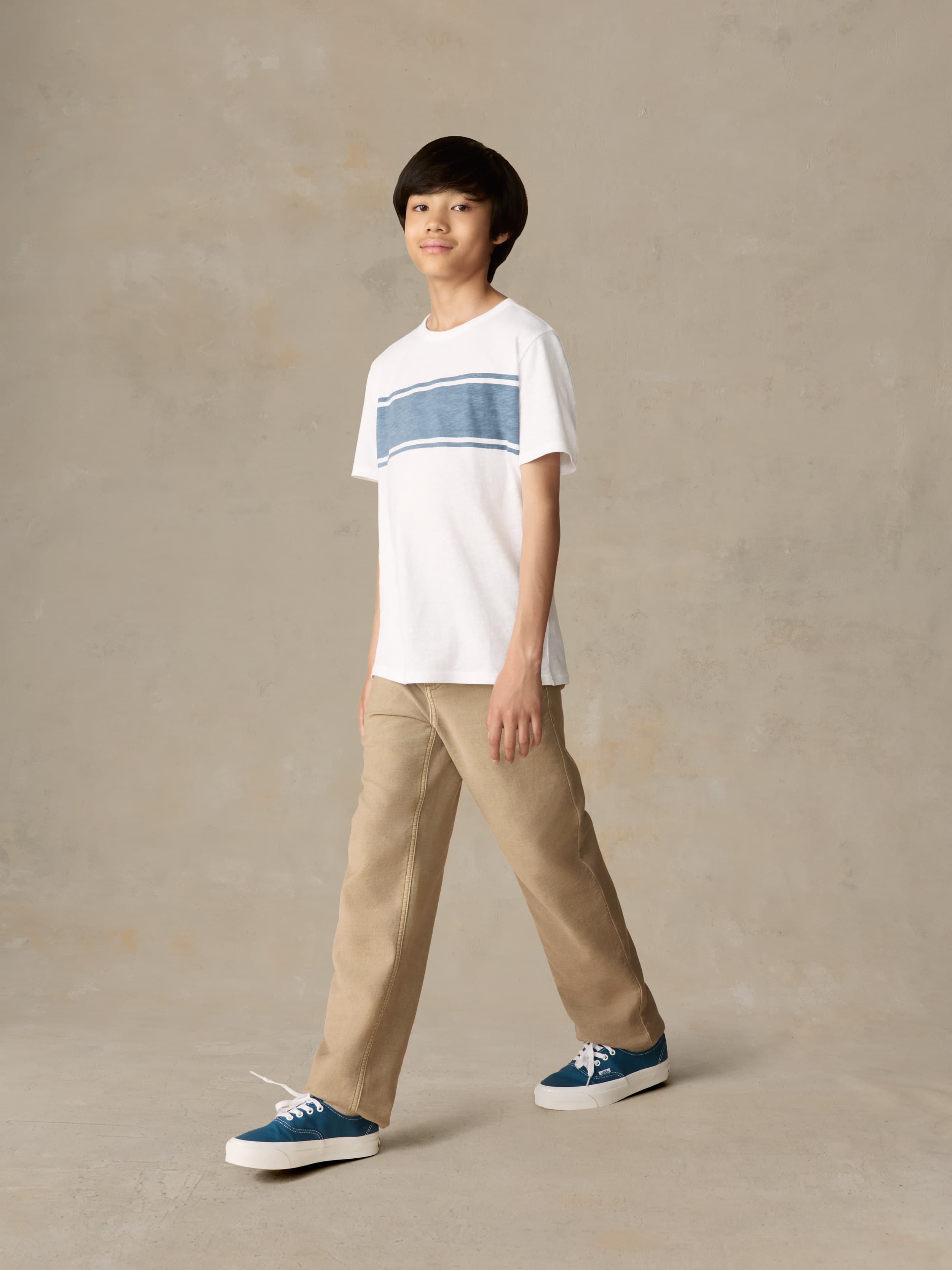 Child wearing a white shirt with blue stripe and beige pants against a beige background