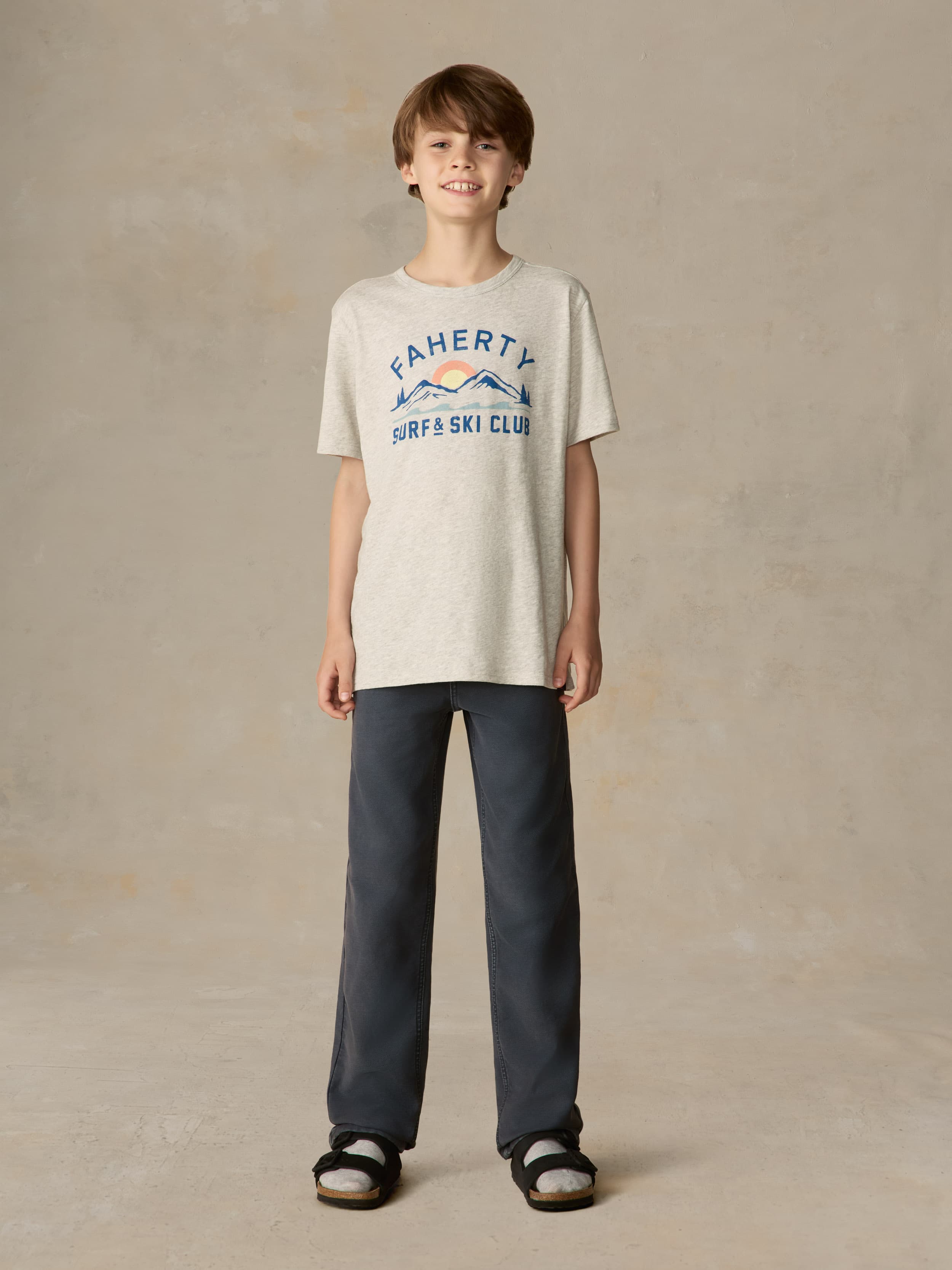 Child wearing a grey surf & ski club tee and black pants against a beige background