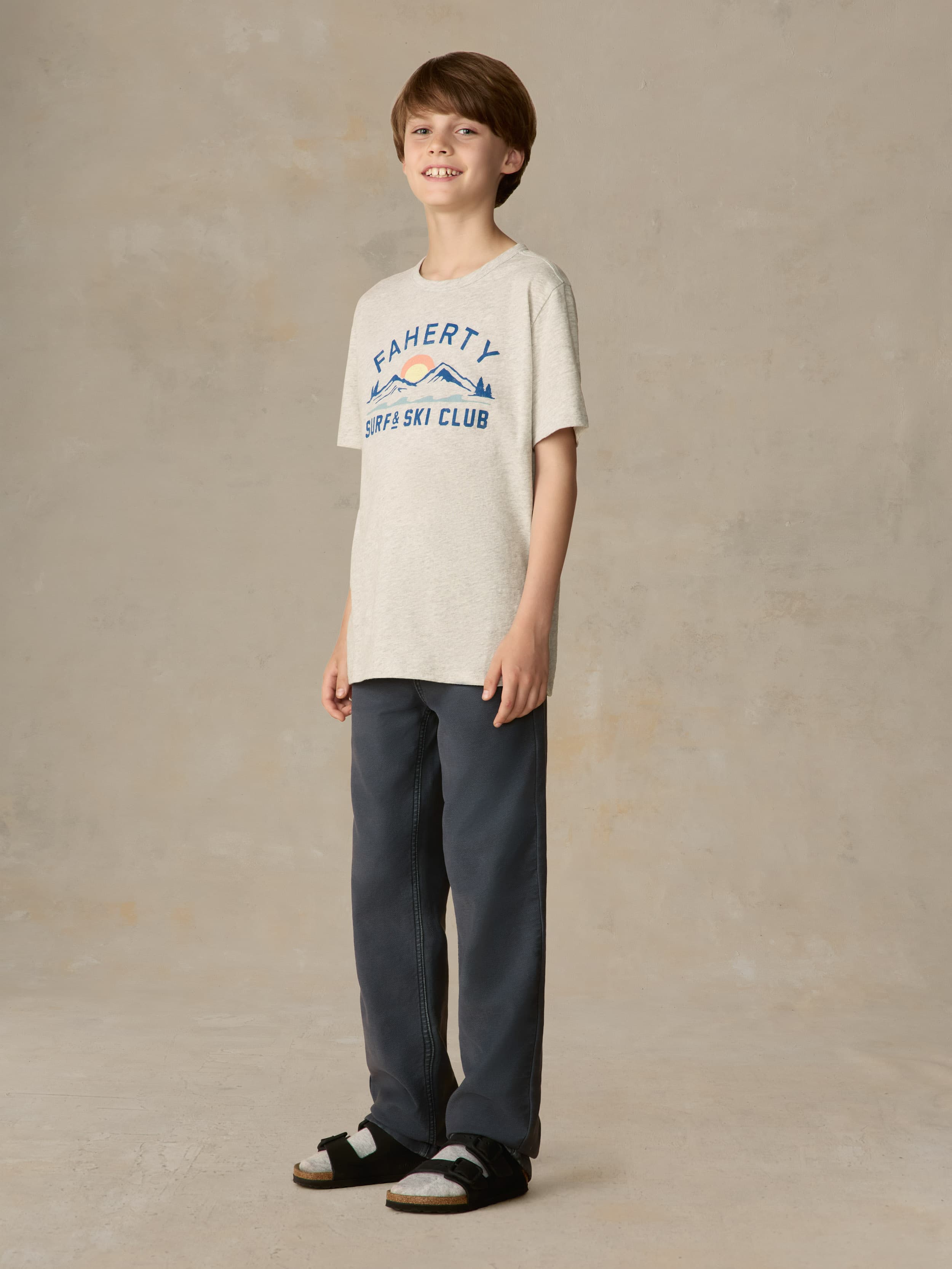 Child wearing a grey surf & ski club tee and black pants against a beige background