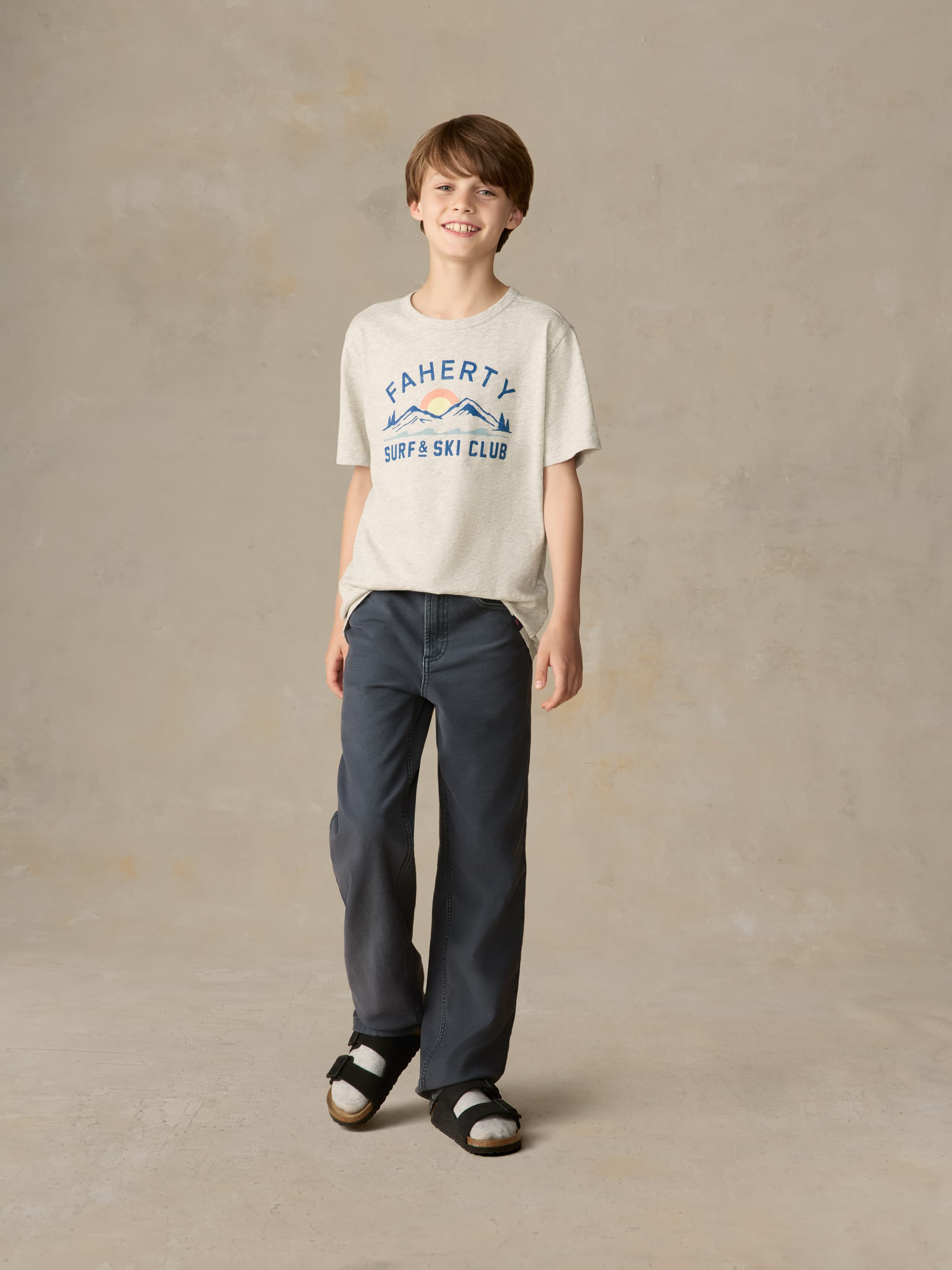 Child wearing a grey surf & ski club tee and black pants against a beige background