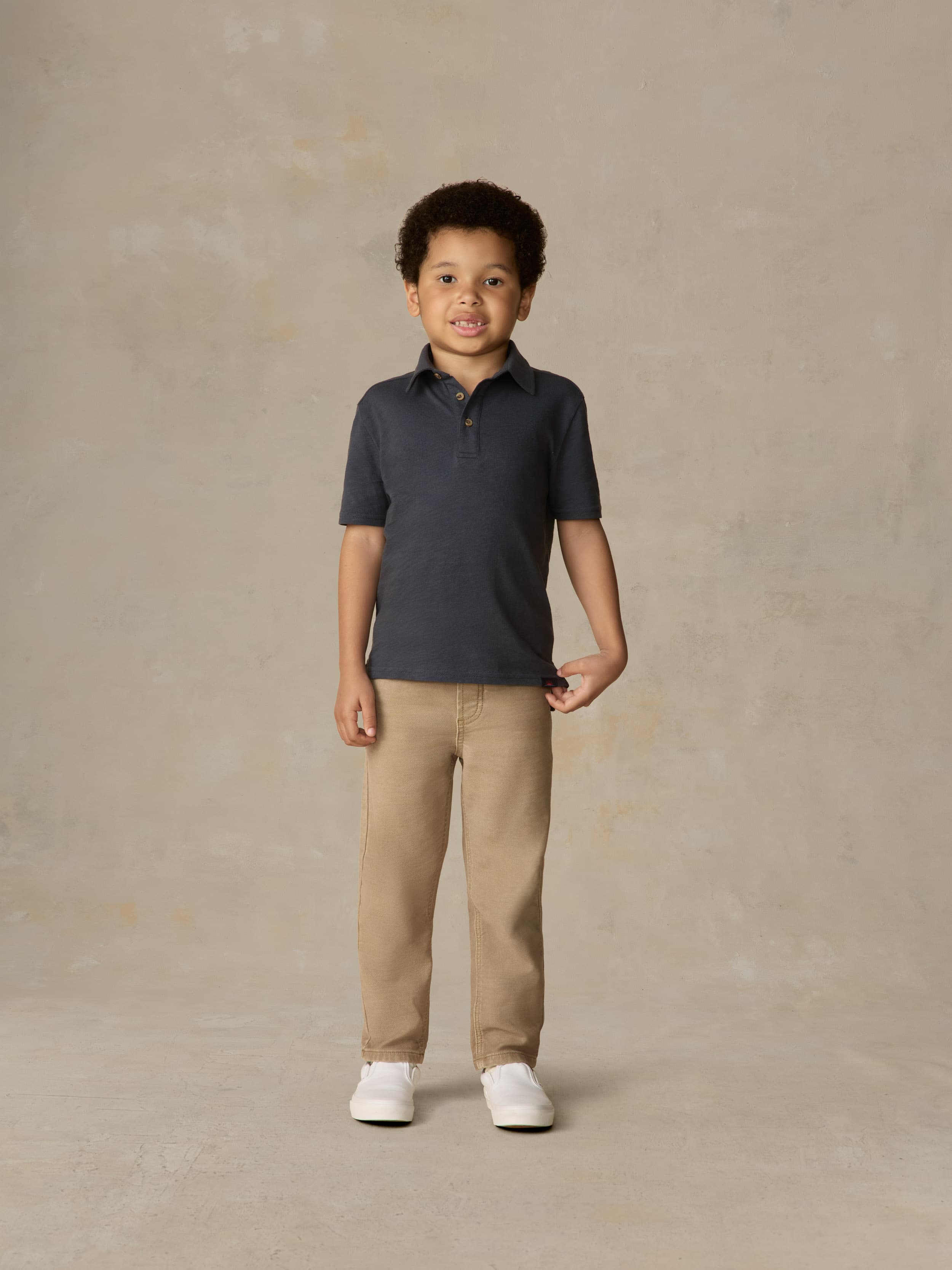 Child wearing a blue polo shirt and beige pants against a beige background