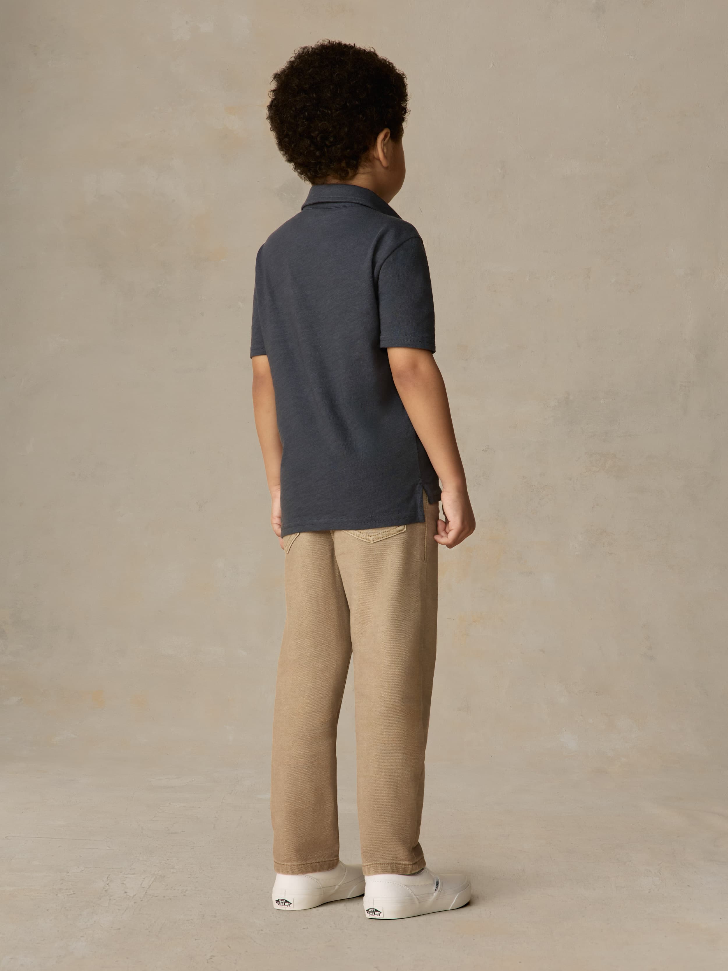 Child wearing a blue polo shirt and beige pants against a beige background