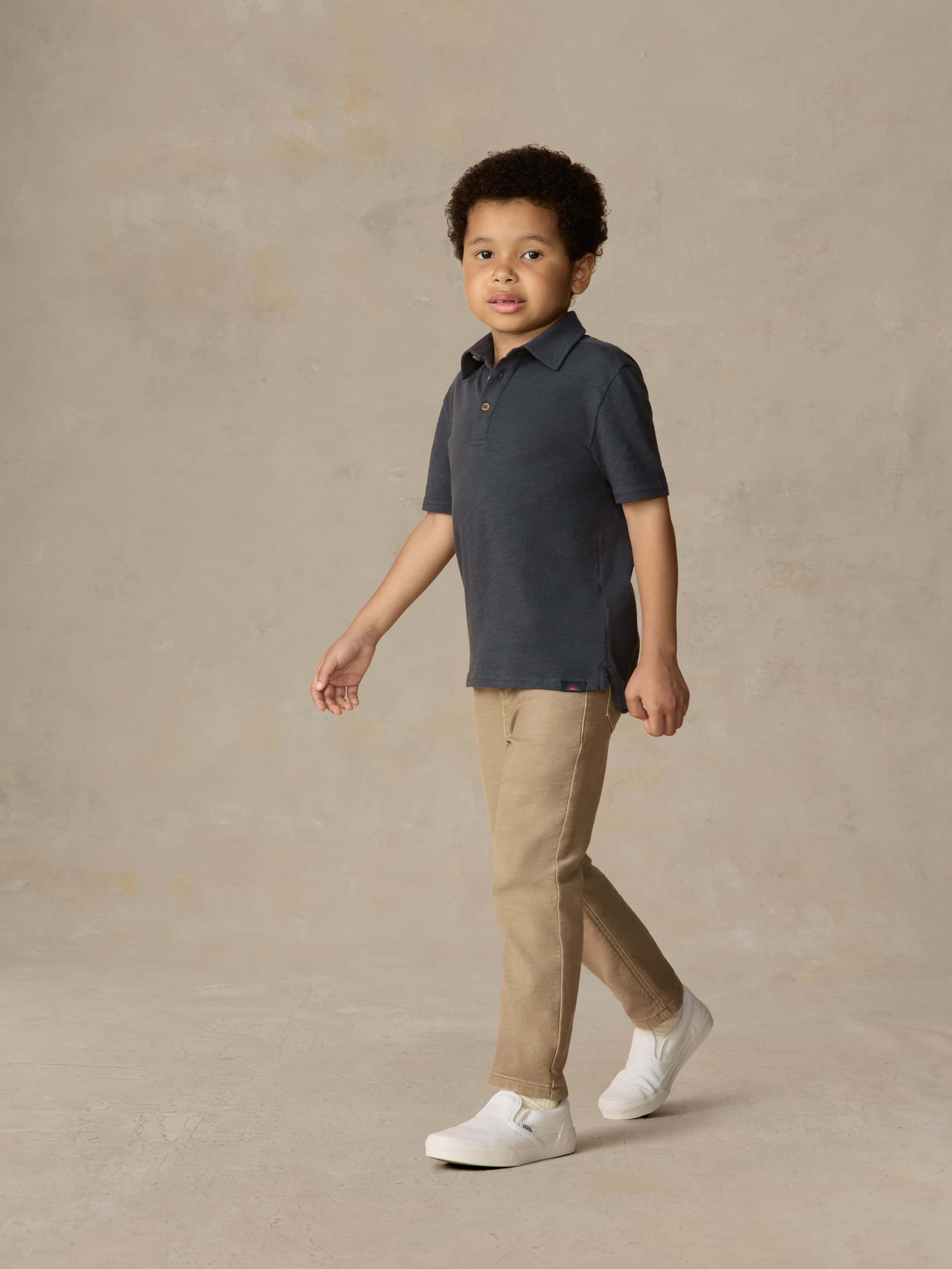 Child wearing a blue polo shirt and beige pants against a beige background
