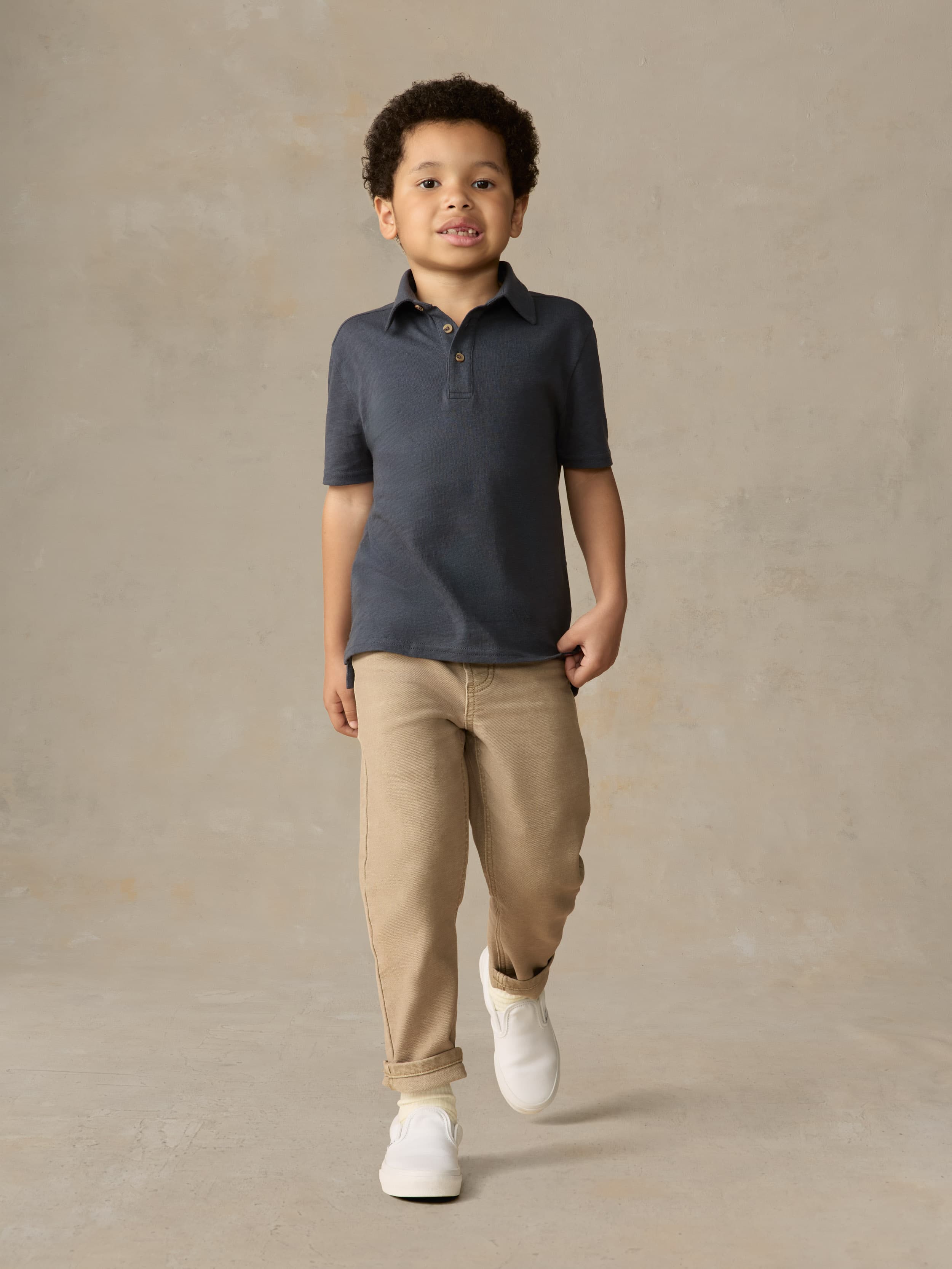 Child wearing a blue polo shirt and beige pants against a beige background