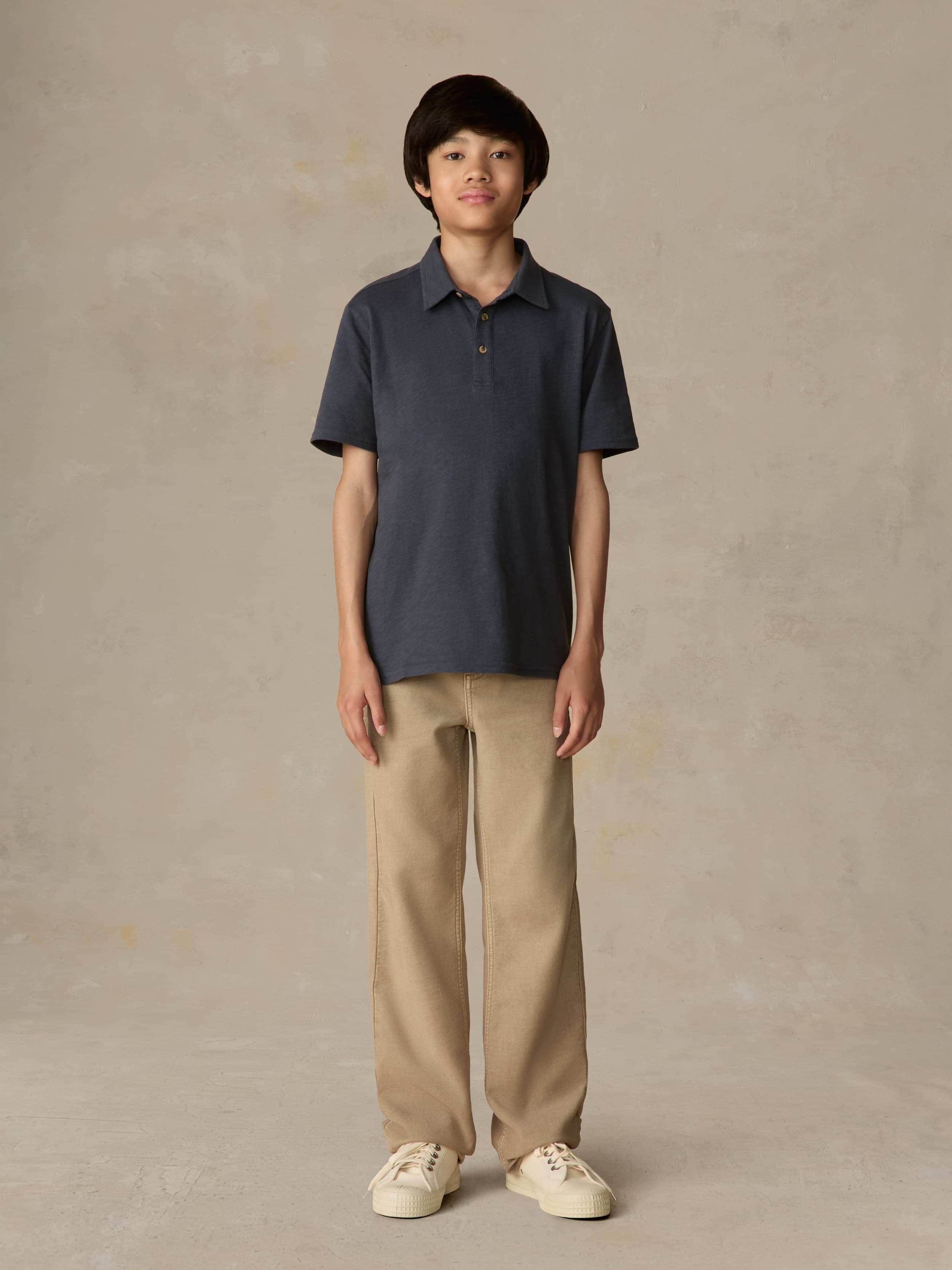 Child wearing a blue polo shirt and beige pants against a beige background