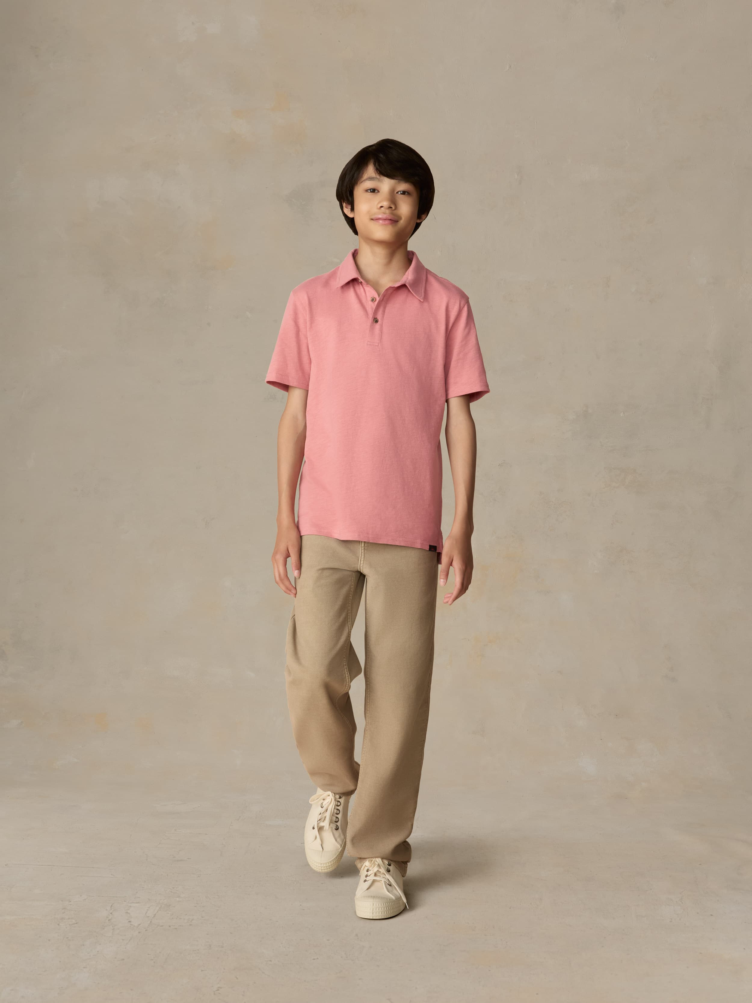 Child wearing a red polo shirt and beige pants against a beige background