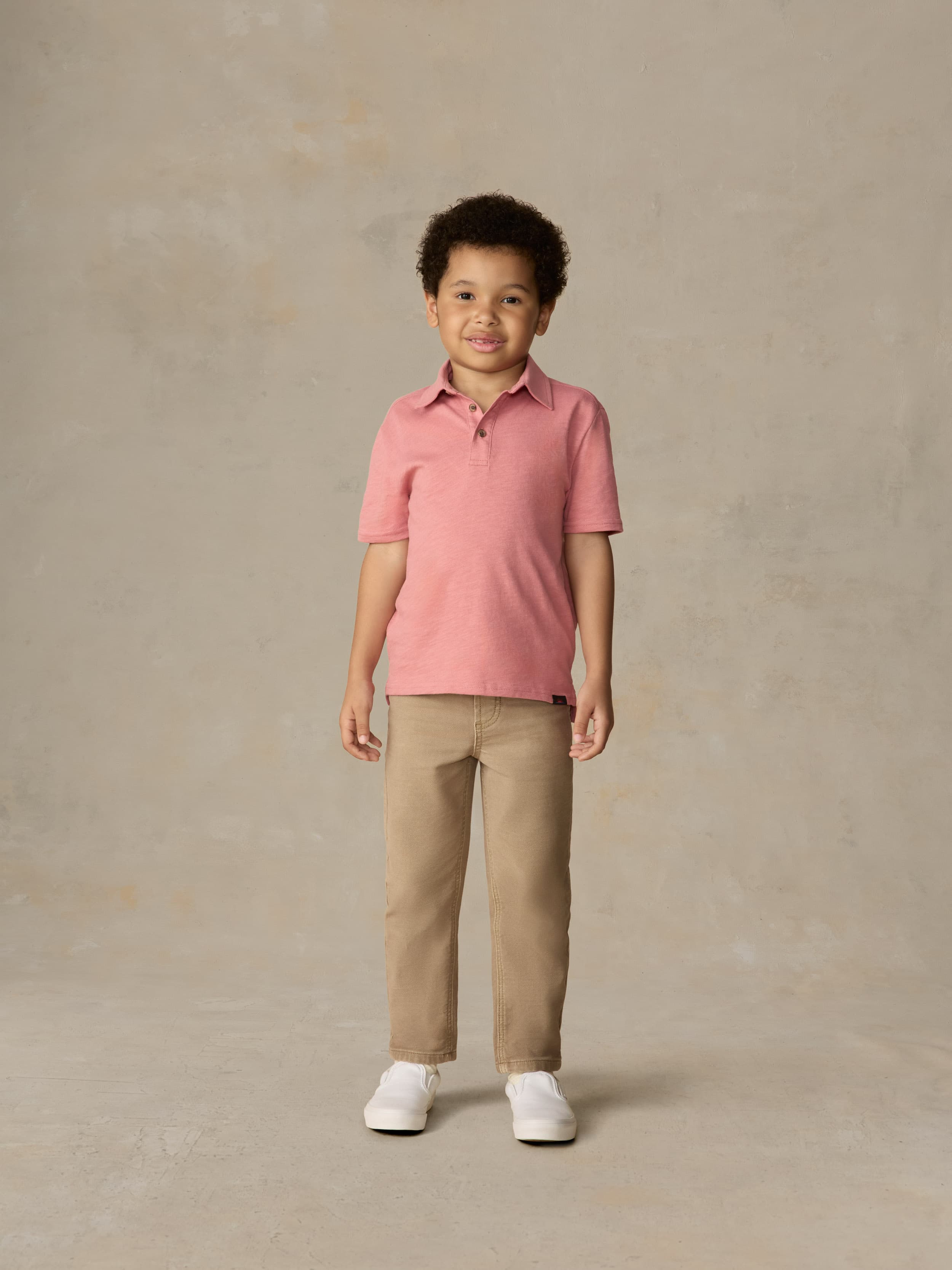 Child wearing a red polo shirt and beige pants against a beige background