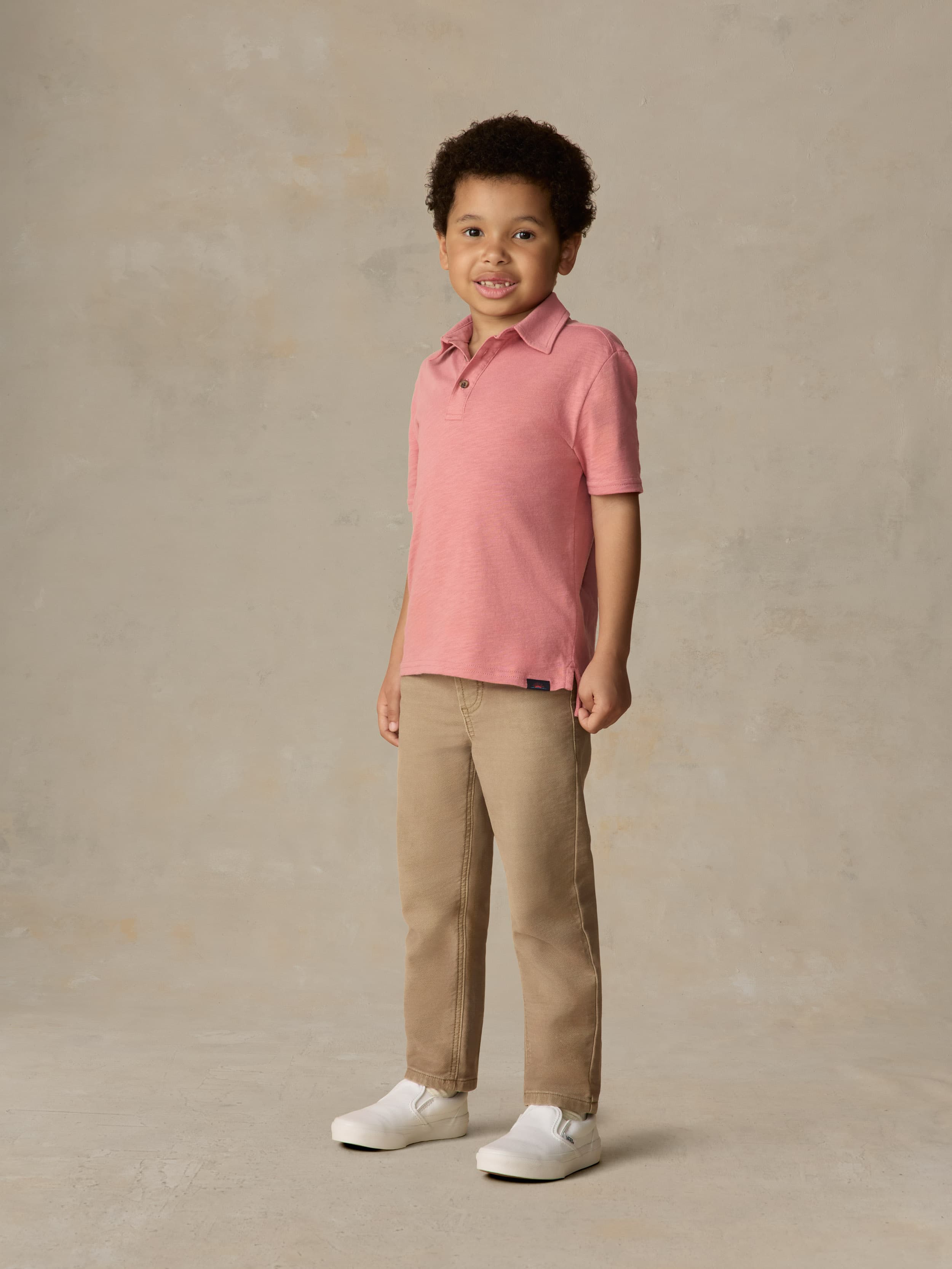Child wearing a red polo shirt and beige pants against a beige background