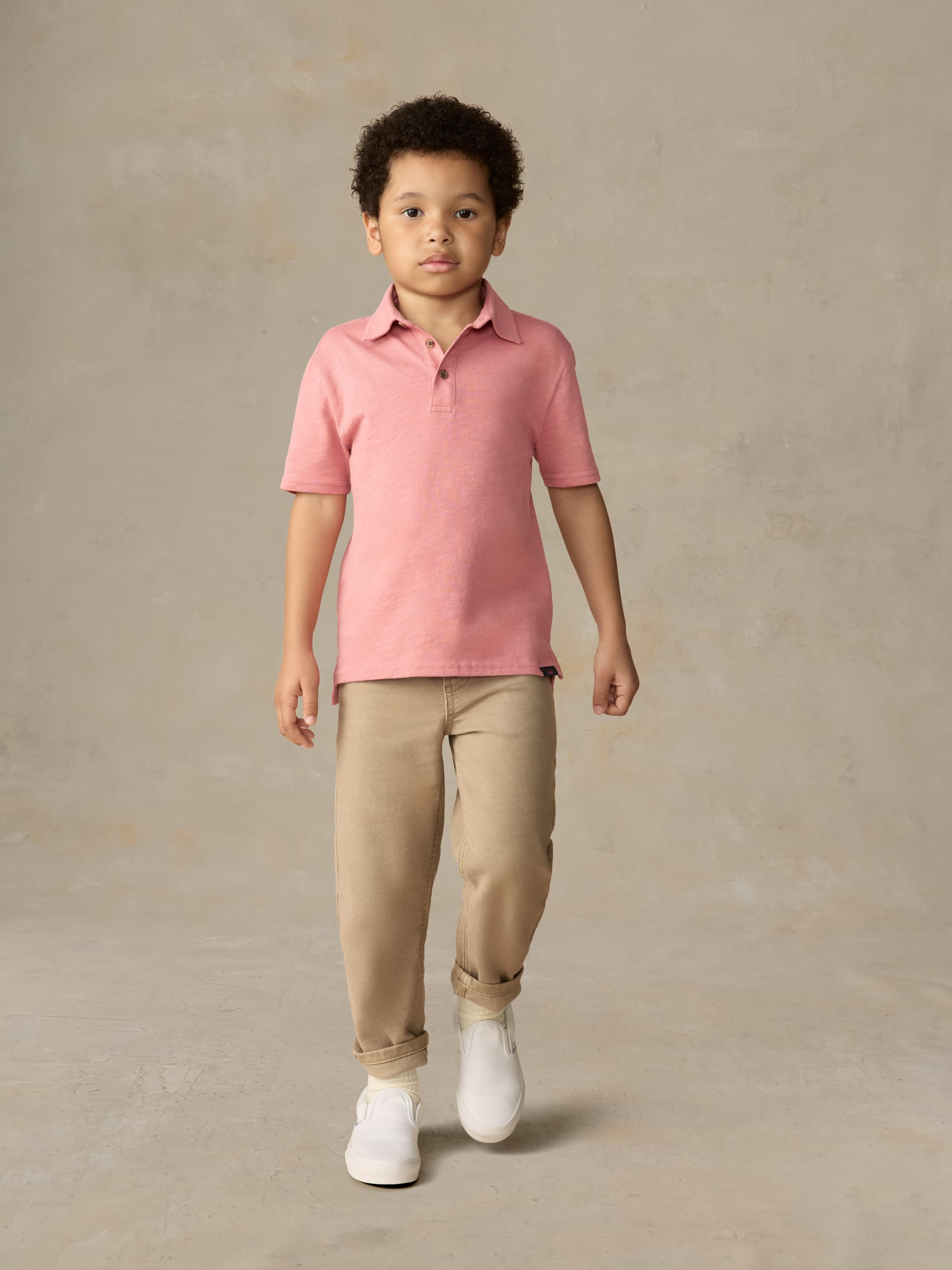 Child wearing a red polo shirt and beige pants against a beige background