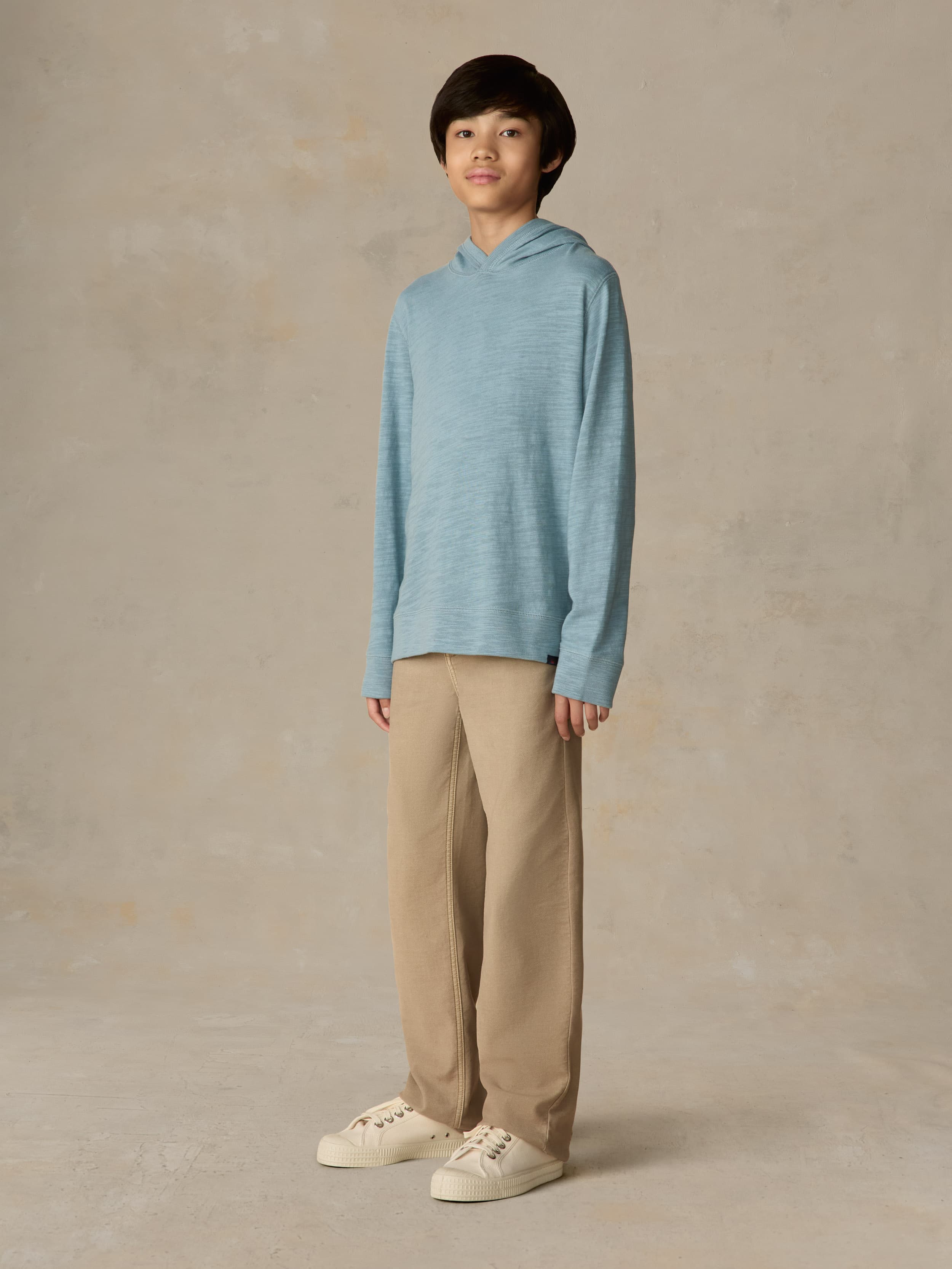 Child wearing a light blue hoodie and beige pants against a beige background