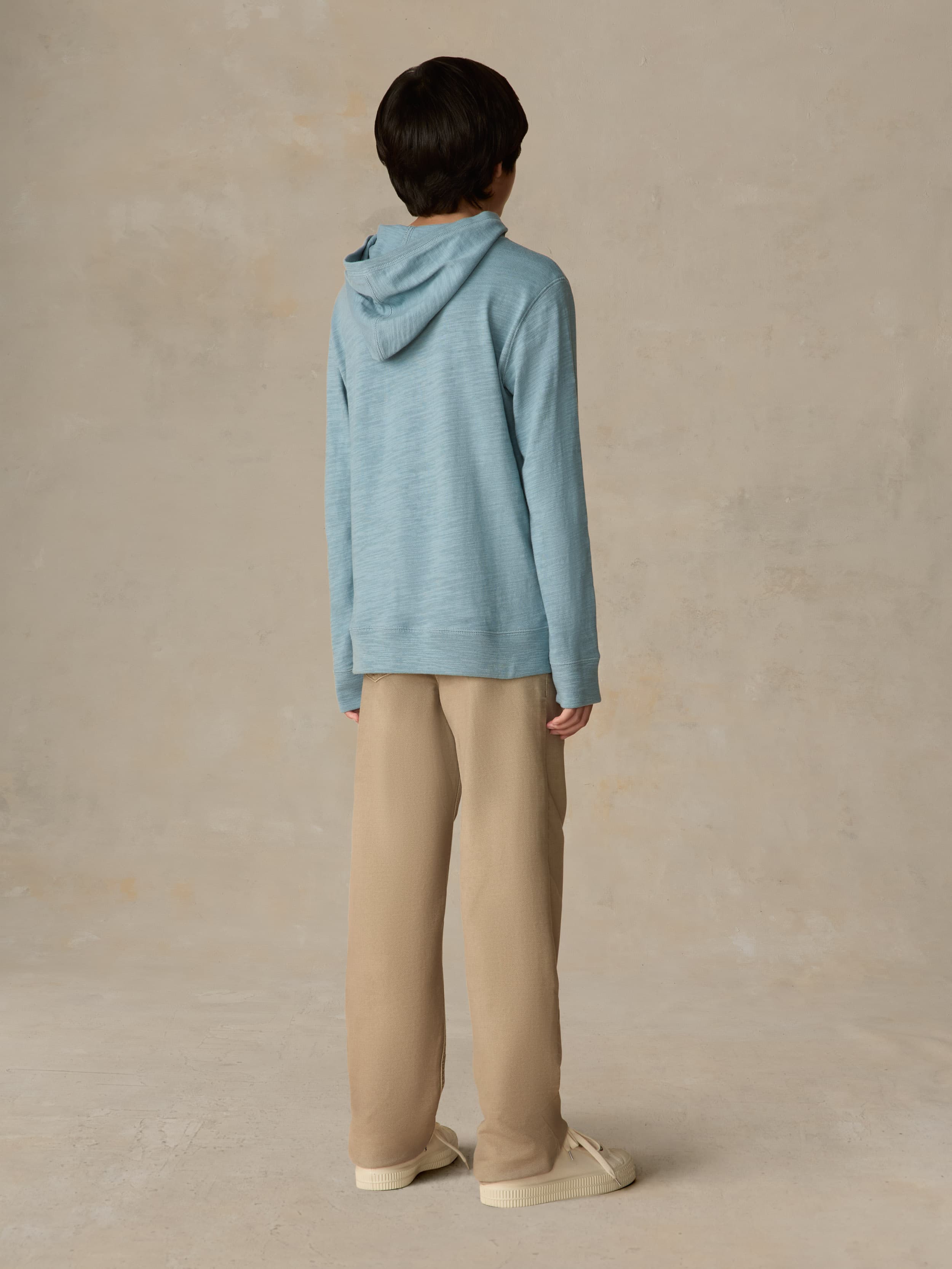 Child wearing a light blue hoodie and beige pants against a beige background