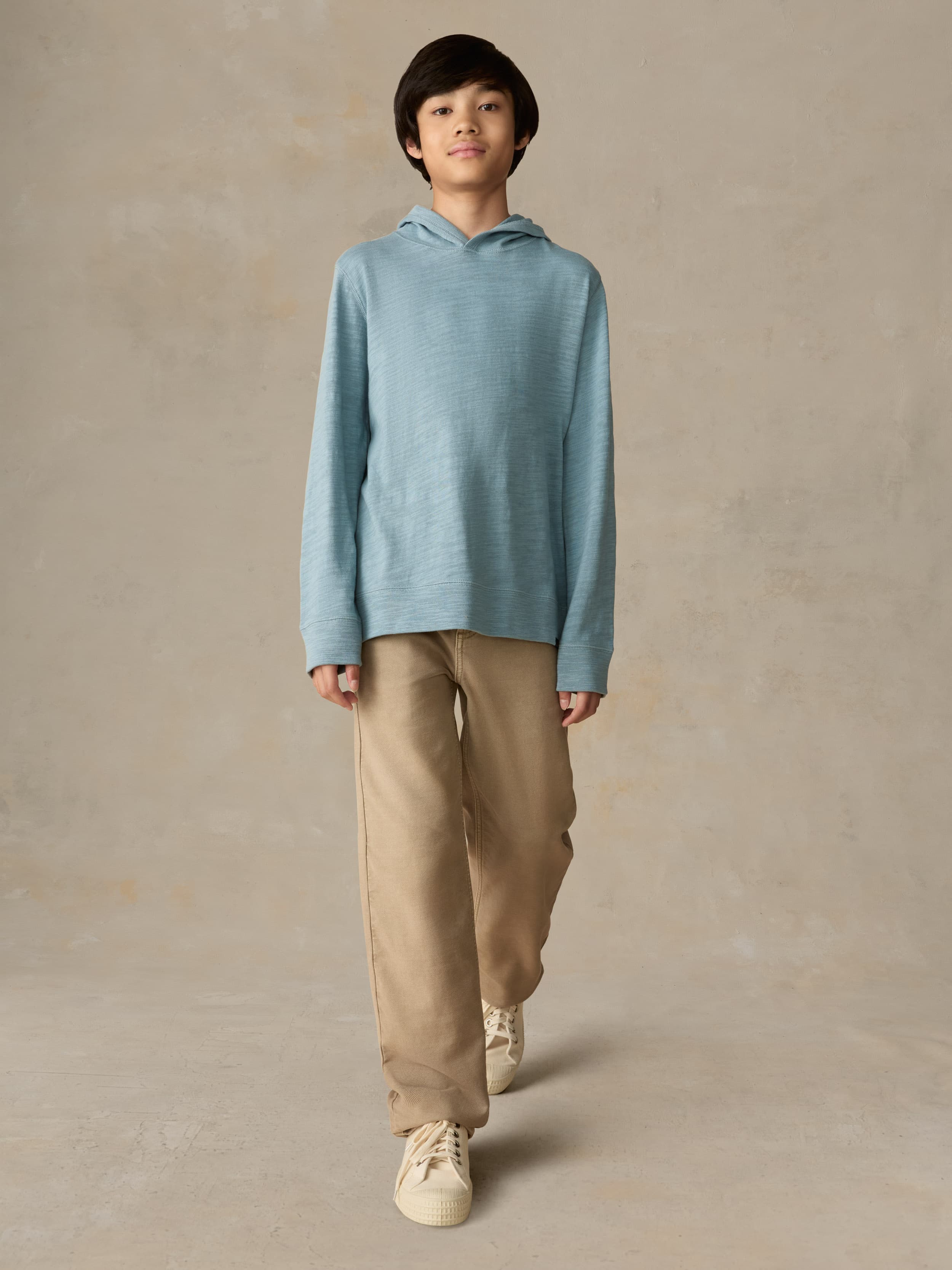 Child wearing a light blue hoodie and beige pants against a beige background