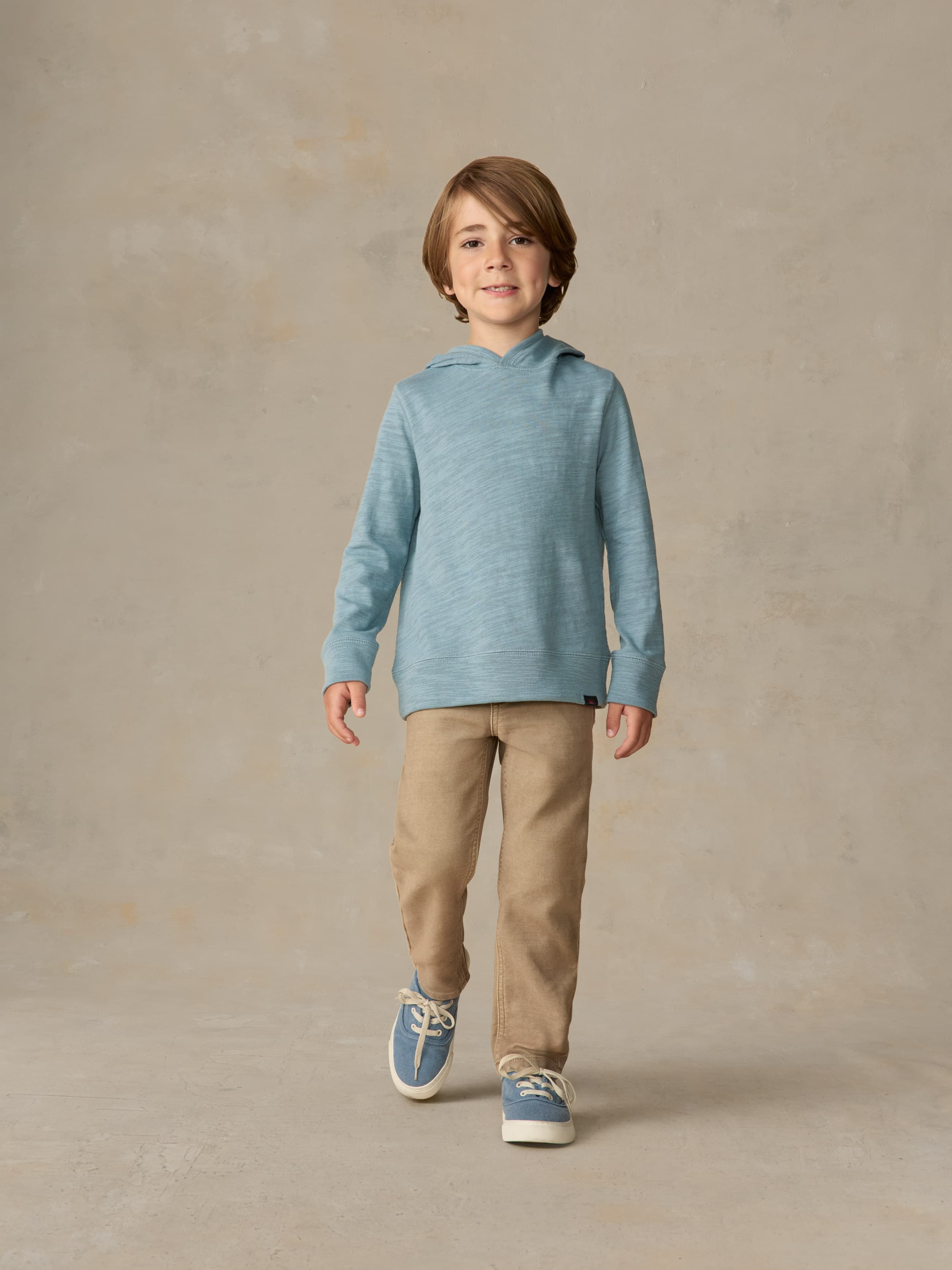 Child wearing a light blue hoodie and beige pants against a beige background